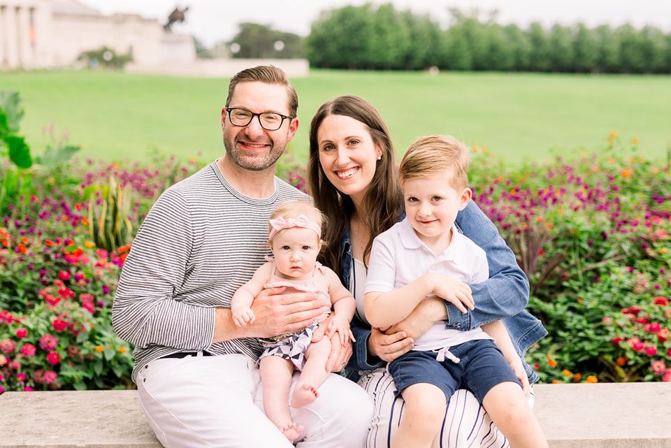 Smiling family of four with infant and toddler boy seated in colorful garden setting.