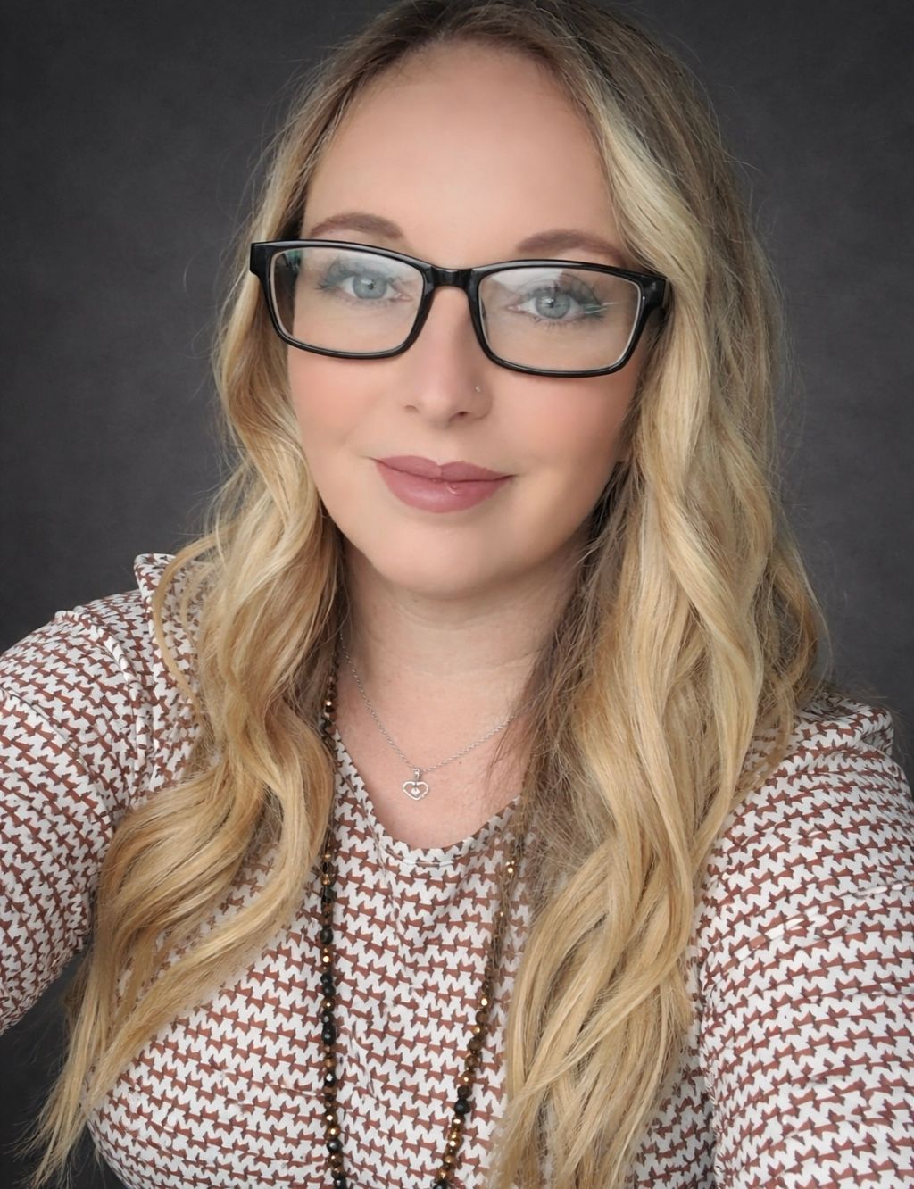 Professional headshot of a blonde woman wearing black glasses and a patterned top with a long beaded necklace.
