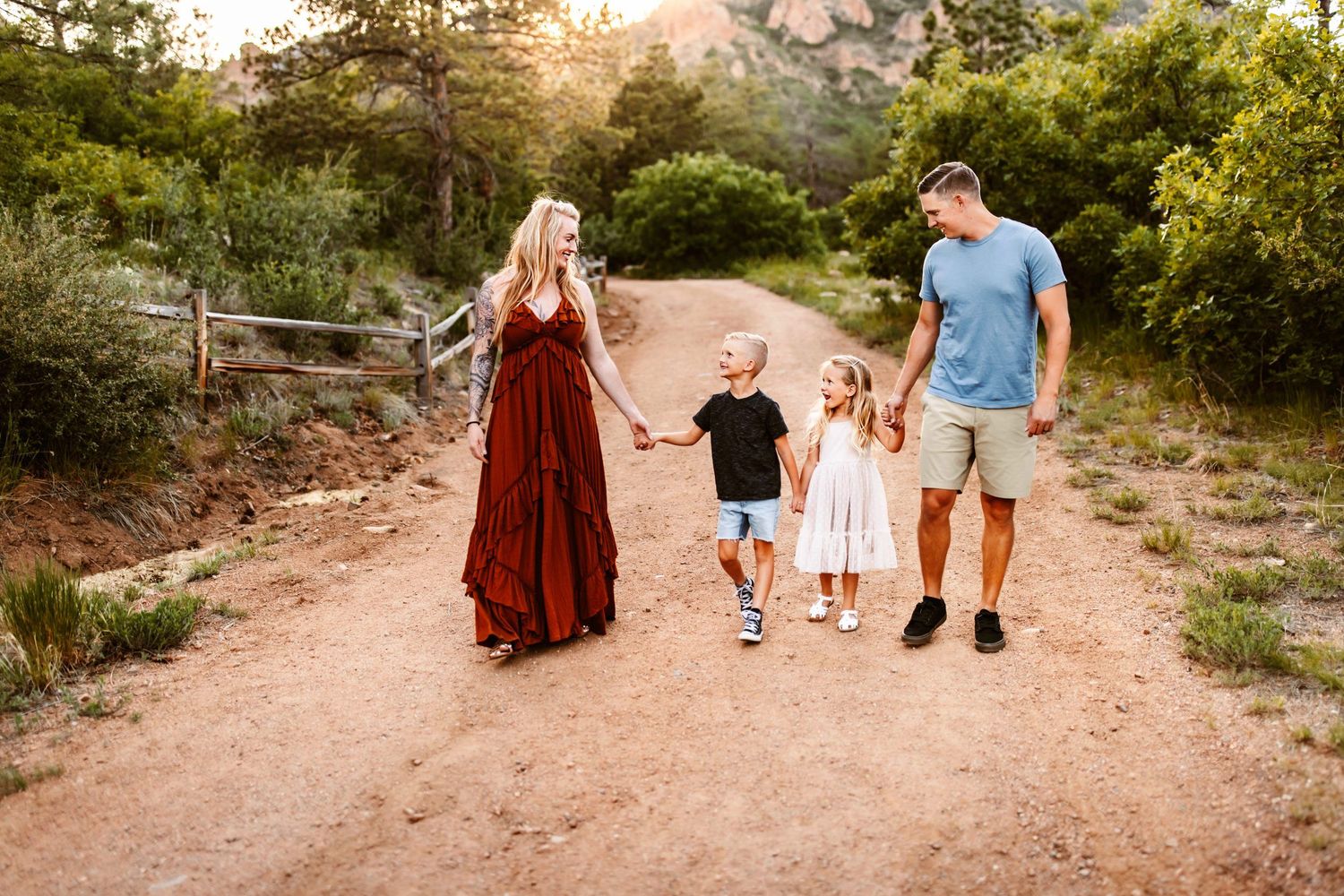 Family of four holding hands walking on rustic dirt path at golden hour sunset.