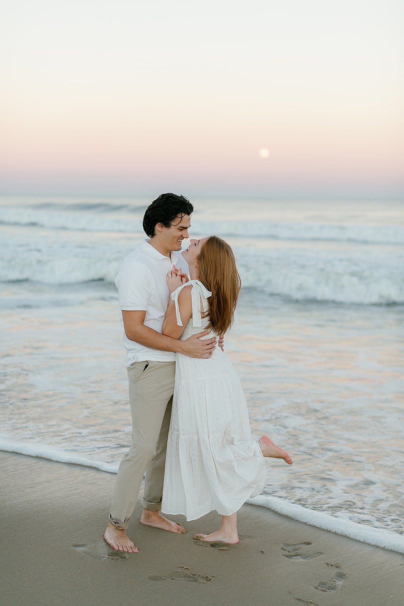Couple embracing barefoot on a serene beach at sunset with gentle waves and a full moon in the sky.