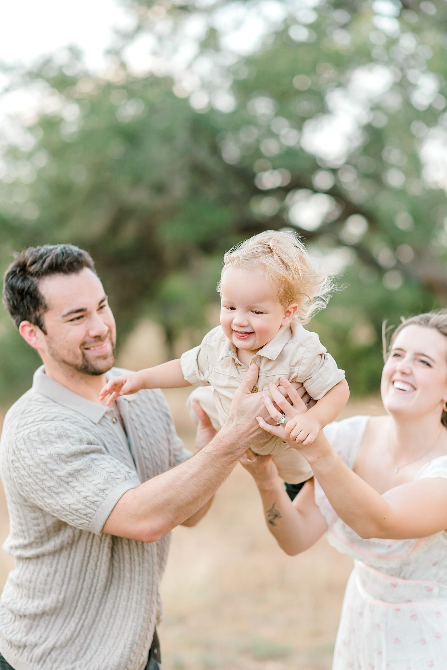 Happy parents lifting their laughing toddler in the air during a sunny outdoor family photo session.