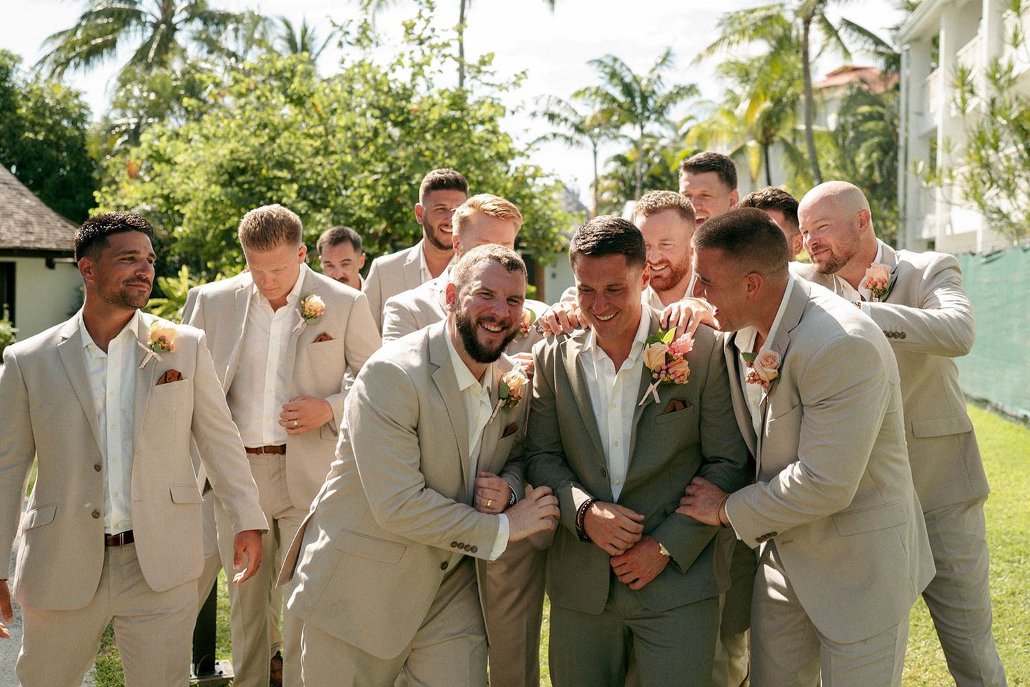 Groom in sage suit surrounded by laughing groomsmen in tan suits outdoors at tropical wedding venue.