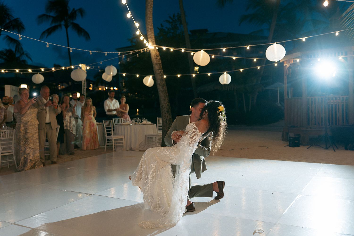 Bride and groom share romantic first dance dip under string lights and lanterns at outdoor beach wedding reception.