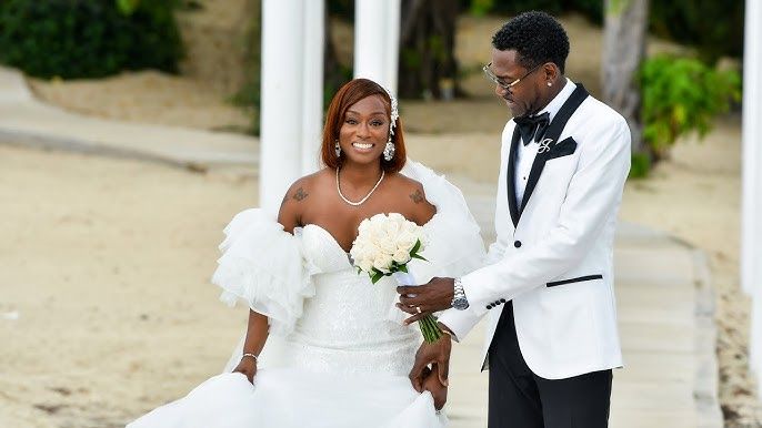 Happy Black couple in wedding attire on beach, bride in white gown with bouquet, groom in white tuxedo jacket.
