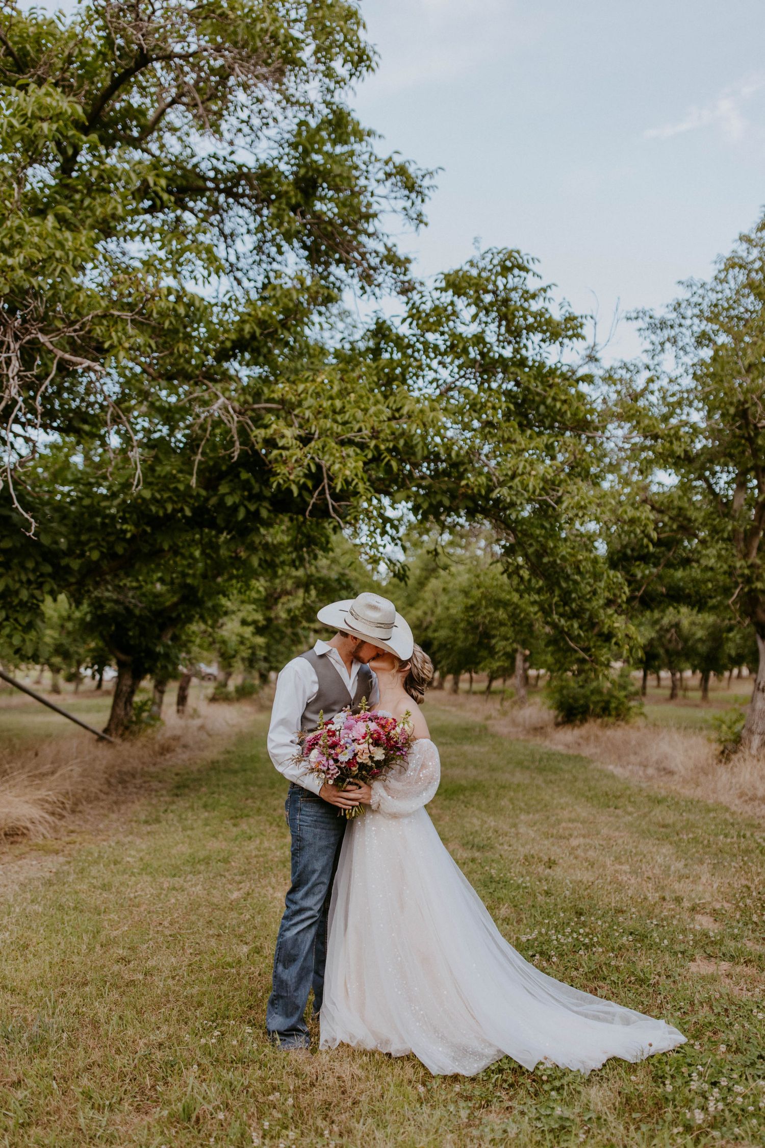 Bride and groom sharing a kiss in an orchard, groom wearing cowboy hat holding a pink wildflower bouquet.