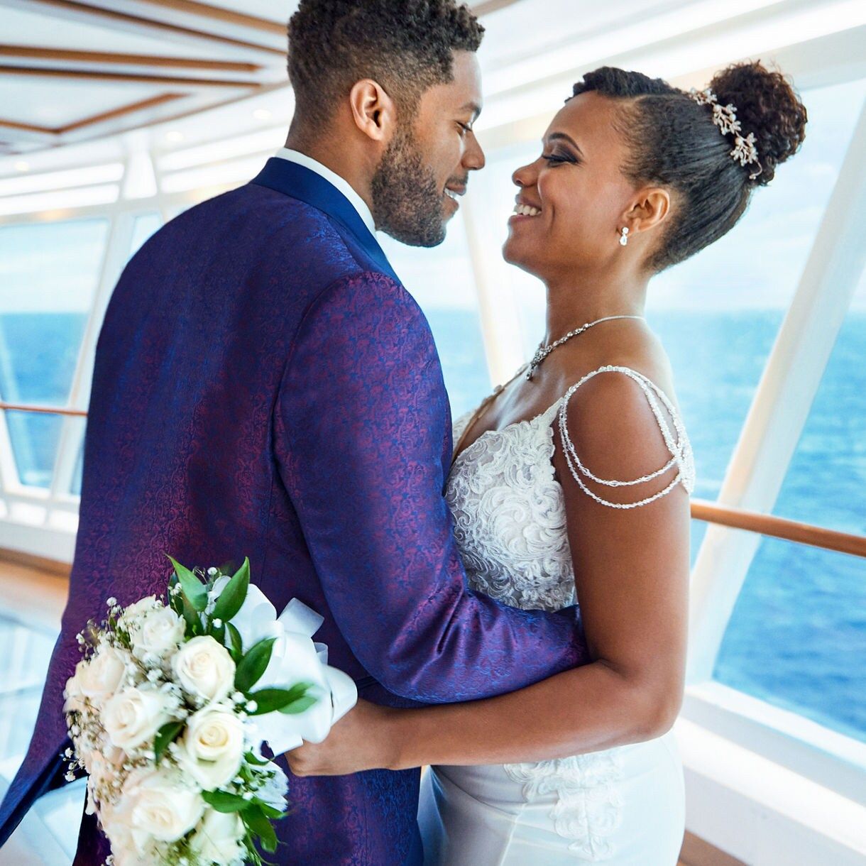 Couple in wedding attire embracing on a cruise ship deck, groom in blue suit holding white bouquet.
