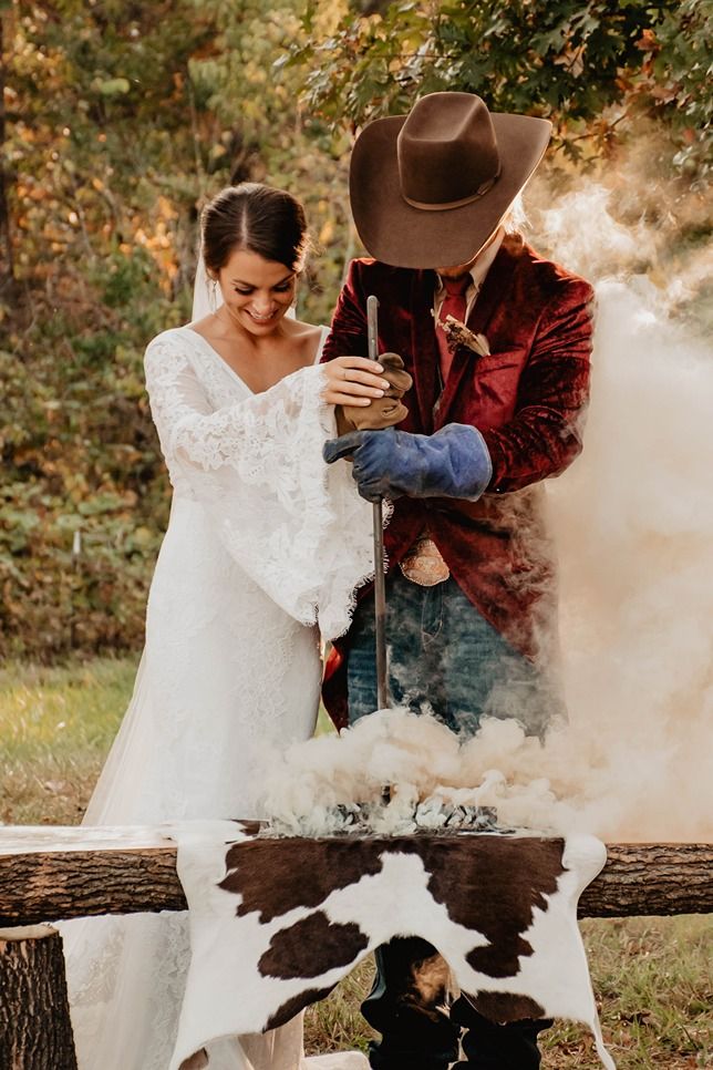 Bride and groom in cowboy hat and velvet blazer performing a sword ceremony with dramatic smoke at rustic outdoor wedding.