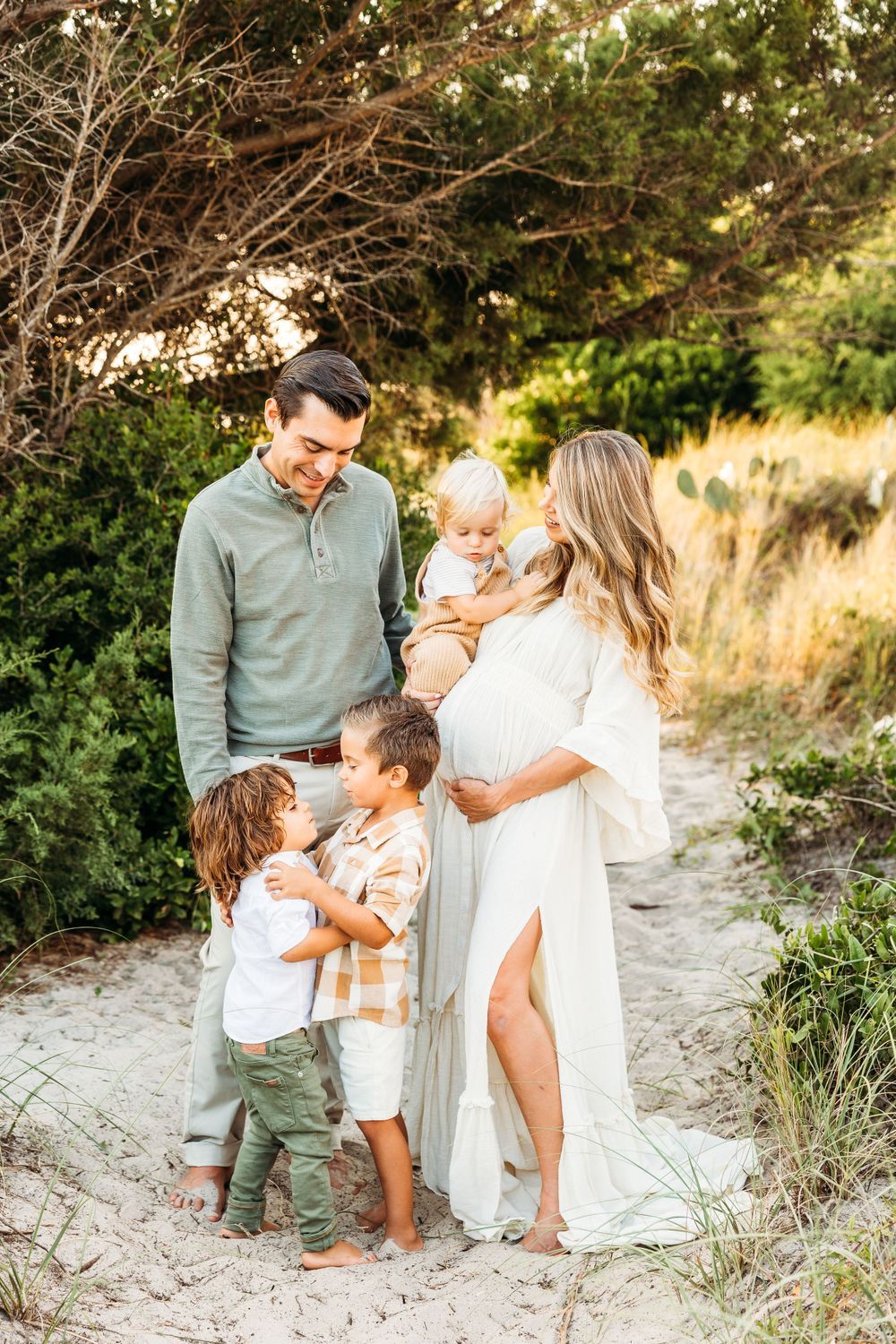 Pregnant mother in white gown with husband and three young children on sandy coastal path.