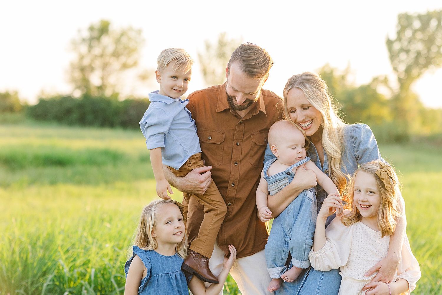 A joyful family of six with four young children posing together in a sunlit green meadow.