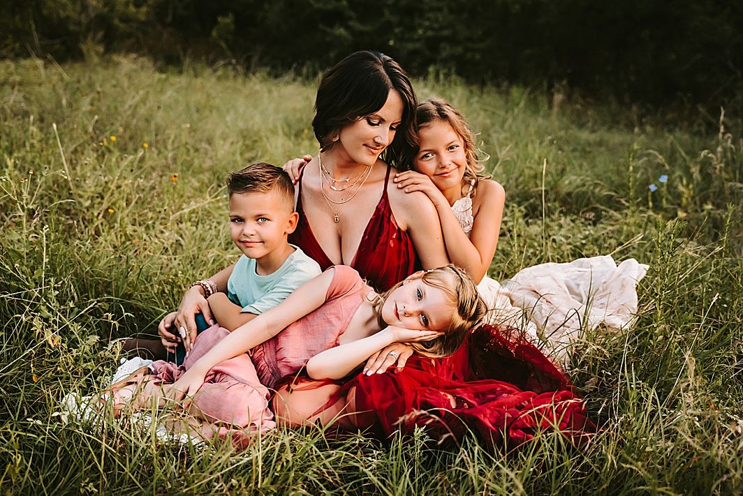 A mother sitting in a grassy field with her three young children during a warm outdoor photo session.
