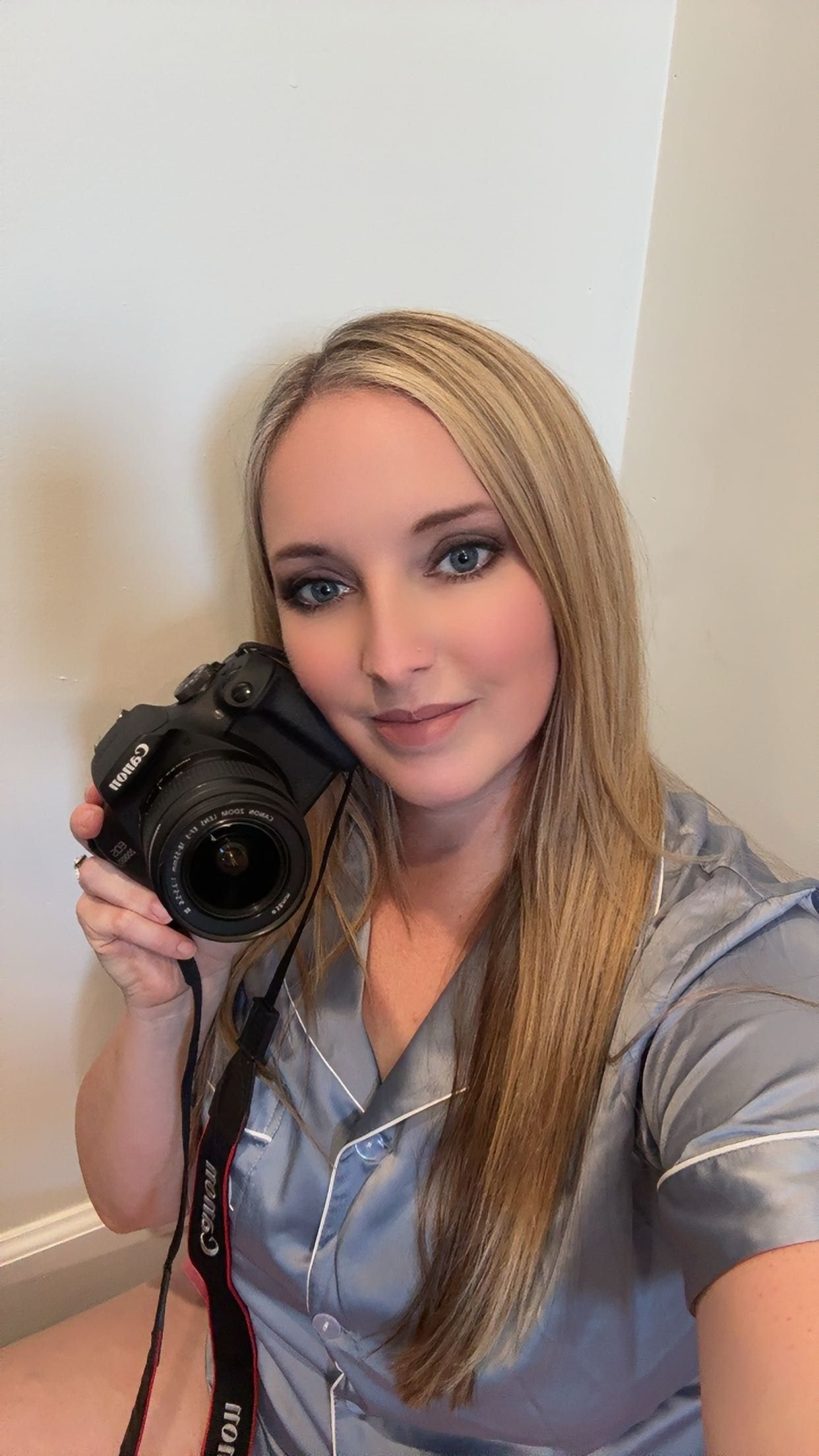 Blonde woman holding a Canon DSLR camera up to her face, wearing a satin gray top, taking a selfie indoors.