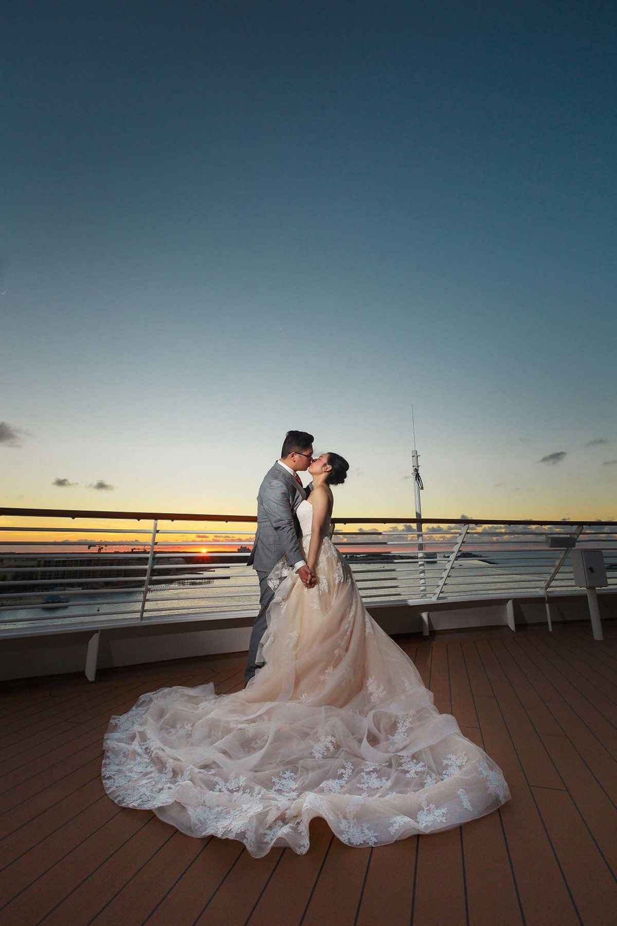Bride and groom share a romantic kiss on a cruise ship deck at sunset, her lace gown trailing elegantly.