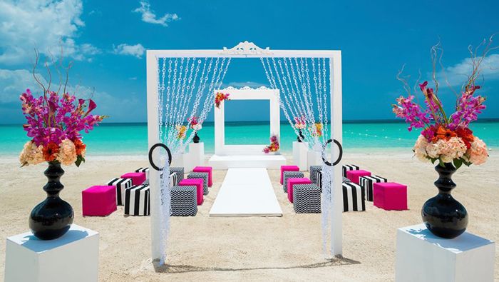 Elegant beach wedding setup with white canopy, pink floral arrangements, and colorful seating on white sand under blue sky.
