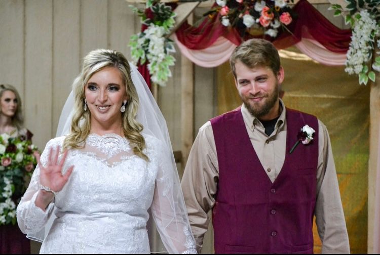 Bride in white lace gown and groom in maroon vest smile at wedding ceremony with floral arch backdrop.