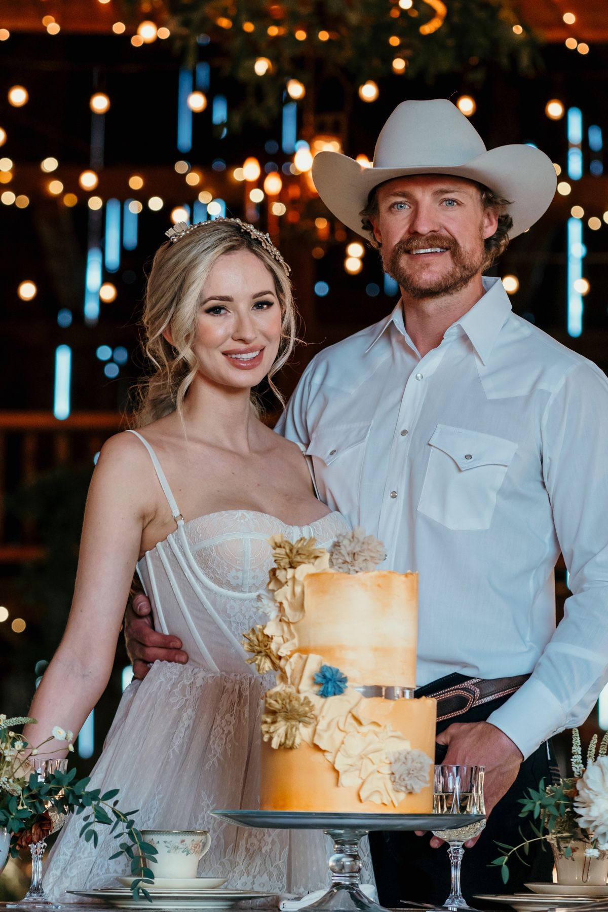 Bride and groom in cowboy hat pose beside a gold tiered wedding cake with floral decorations under warm string lights.