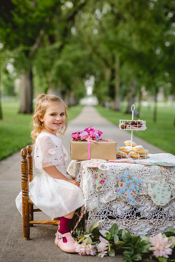Young girl in white dress sitting at outdoor tea party table with treats and flowers in park.