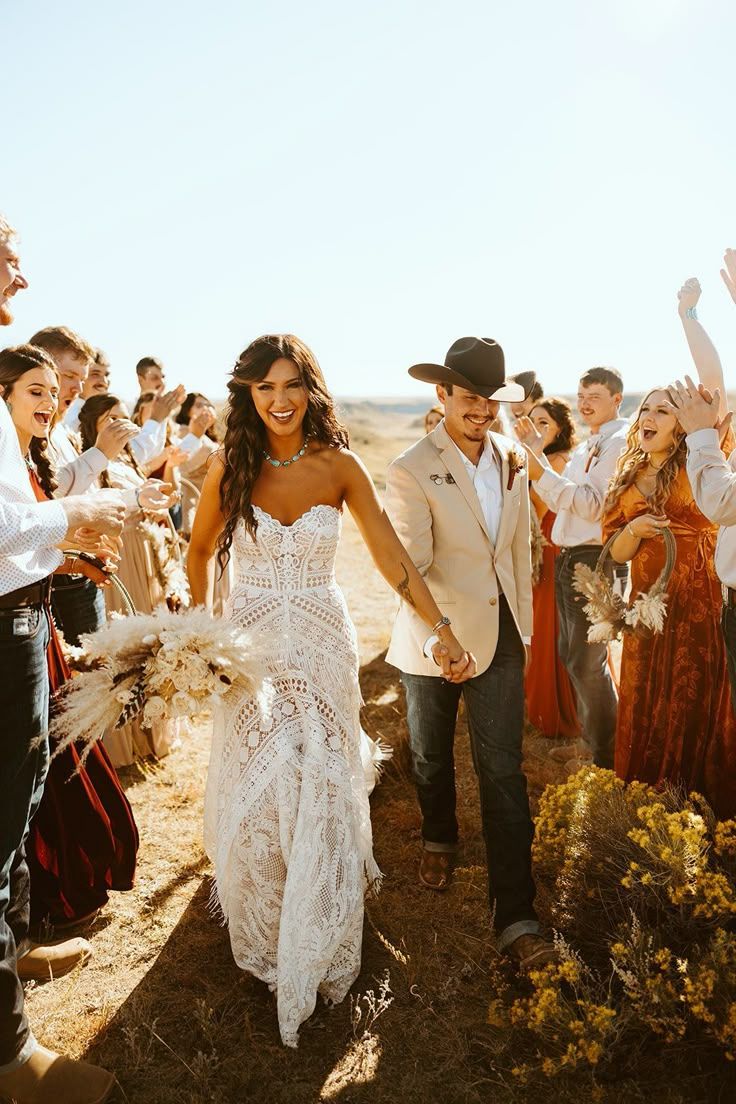 Boho bride in lace gown and groom in cowboy hat walk through cheering guests at outdoor western wedding.