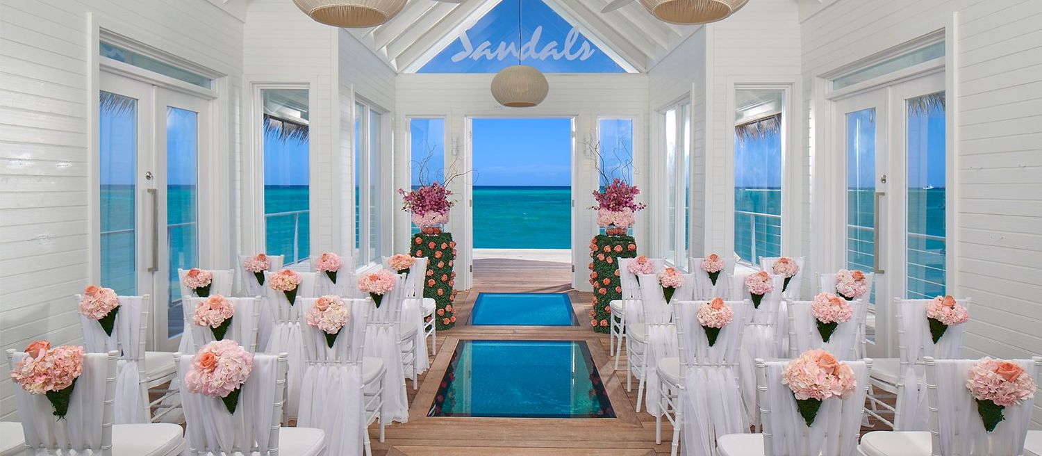 Elegant Sandals resort wedding chapel with white chairs, pink roses, and stunning ocean views through floor-to-ceiling windows.
