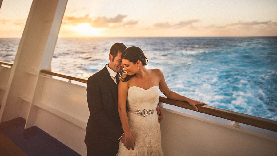 Newlywed couple embracing on cruise ship deck at sunset, bride in strapless lace gown, groom in black tuxedo.