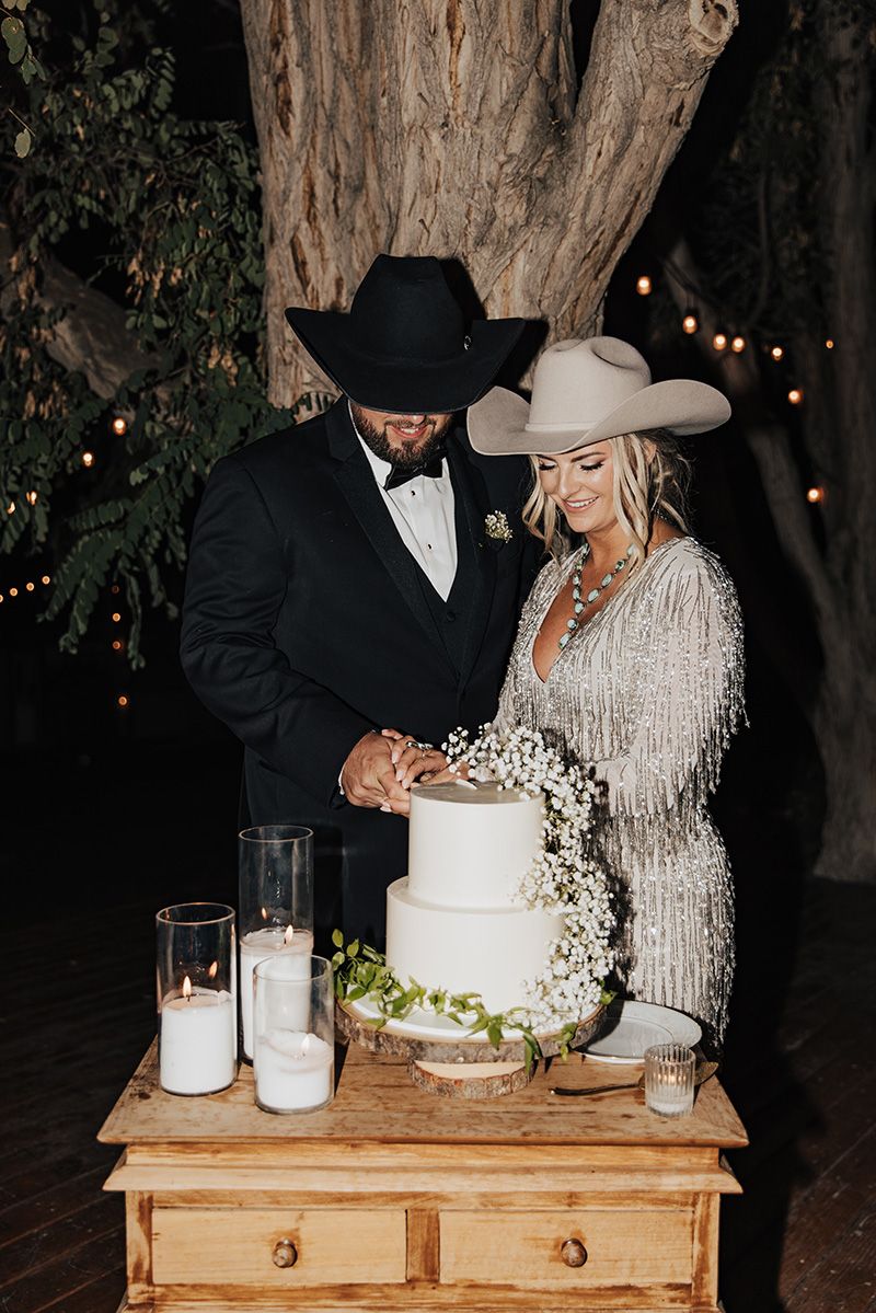Couple in cowboy hats cutting white wedding cake outdoors at night, groom in tuxedo, bride in silver fringe dress.