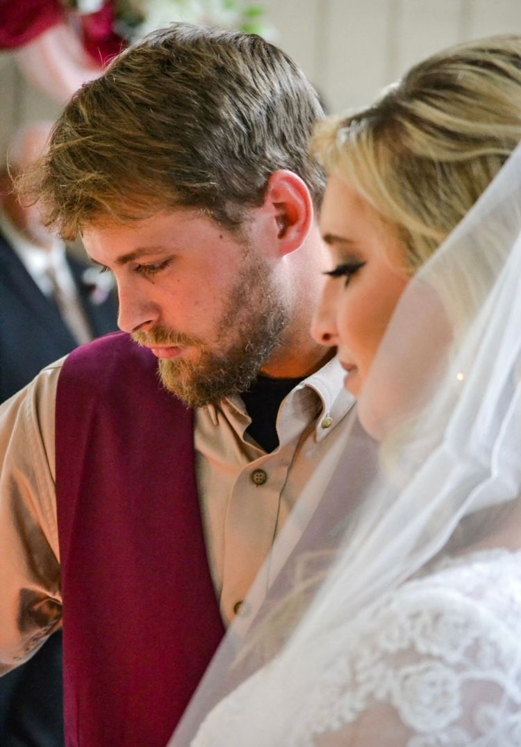 A bride in a lace veil and groom in a burgundy vest bow their heads in prayer during a wedding ceremony.