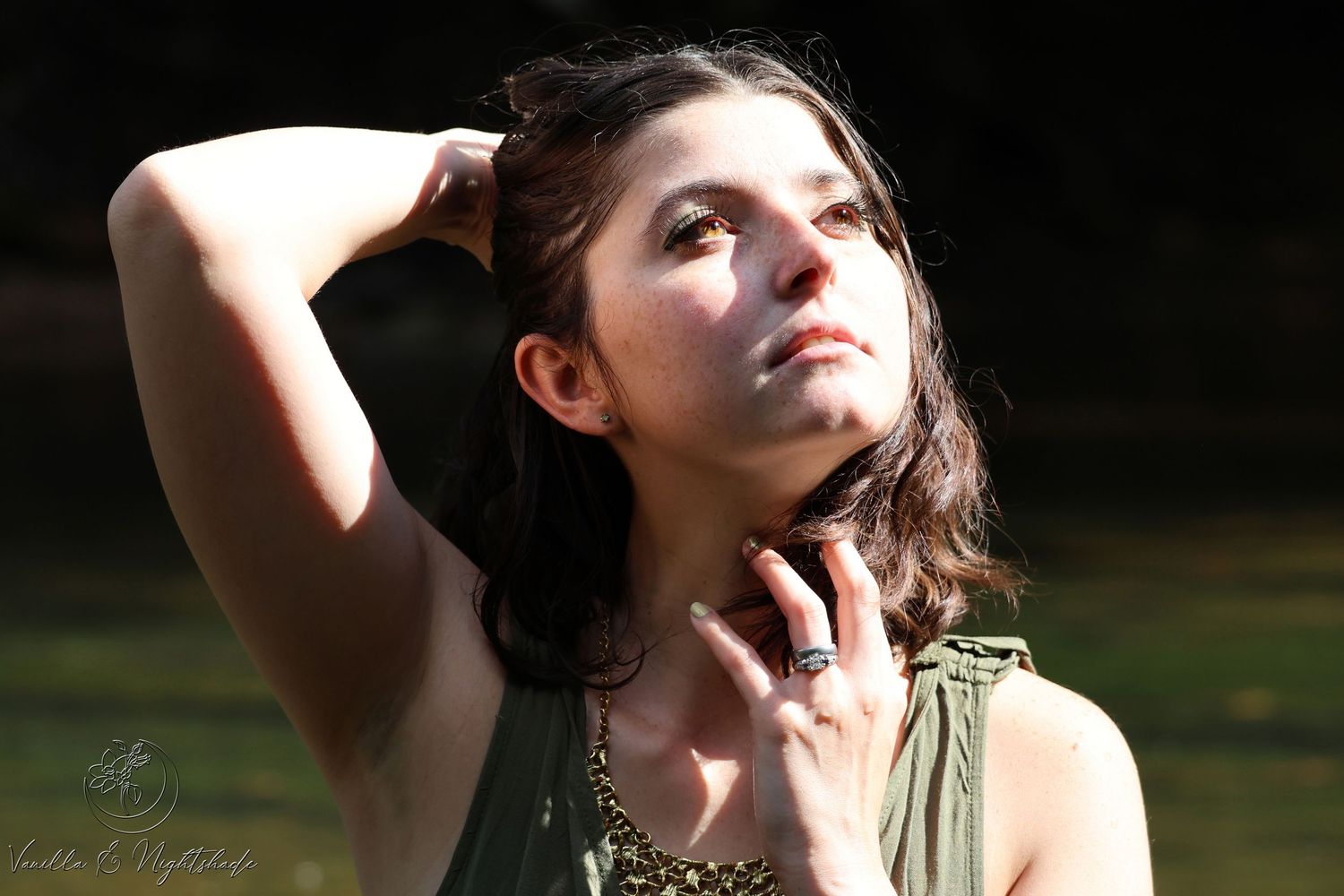 Young woman in olive dress gazes upward, hand in dark hair, sunlight filtering through trees outdoors.