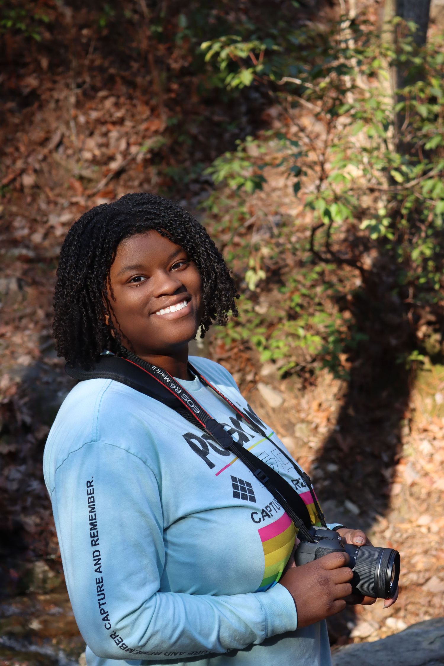 Smiling young Black woman holding a DSLR camera outdoors in autumn forest setting.
