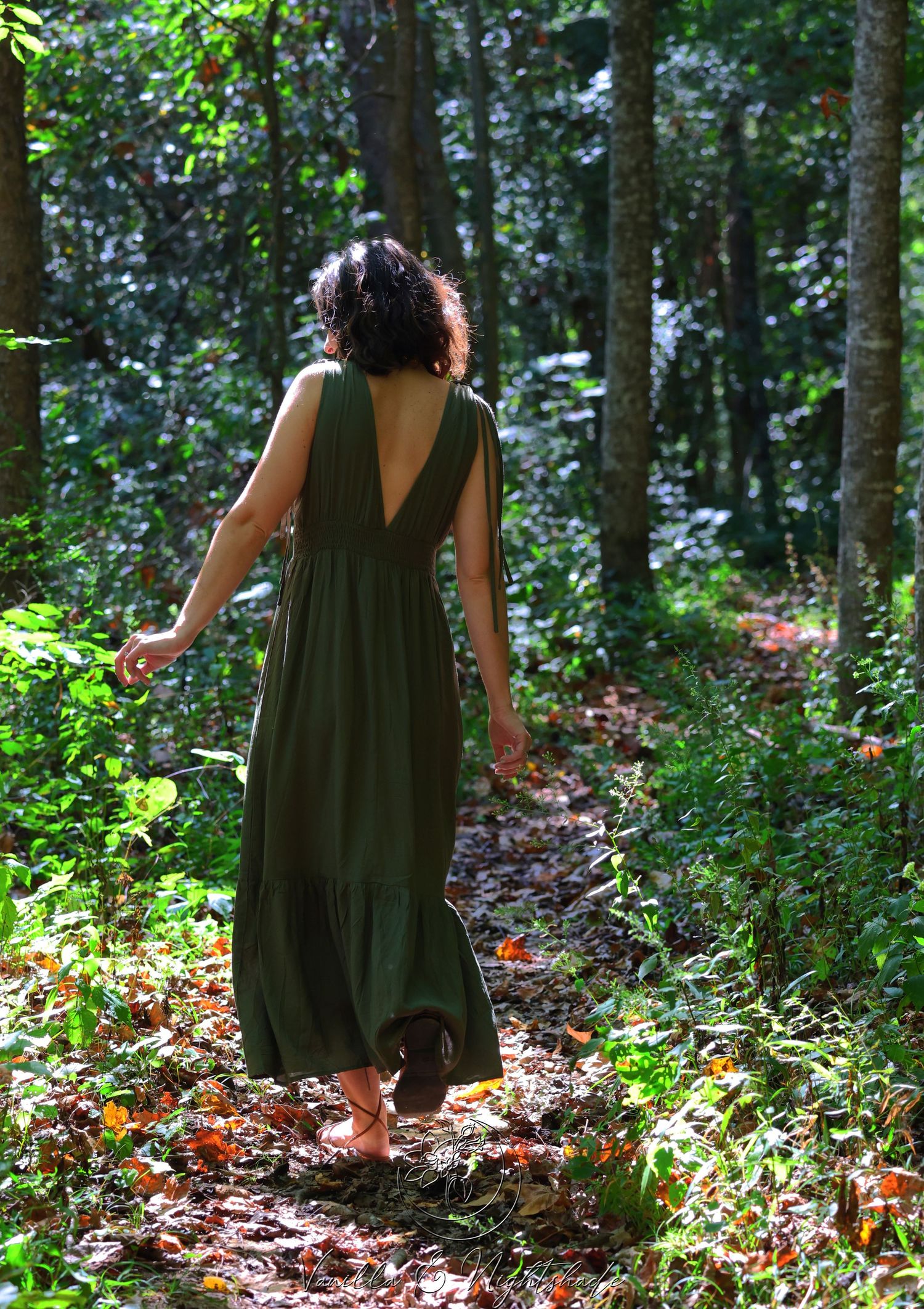 Barefoot woman in long olive green dress walking along a sunlit forest trail.