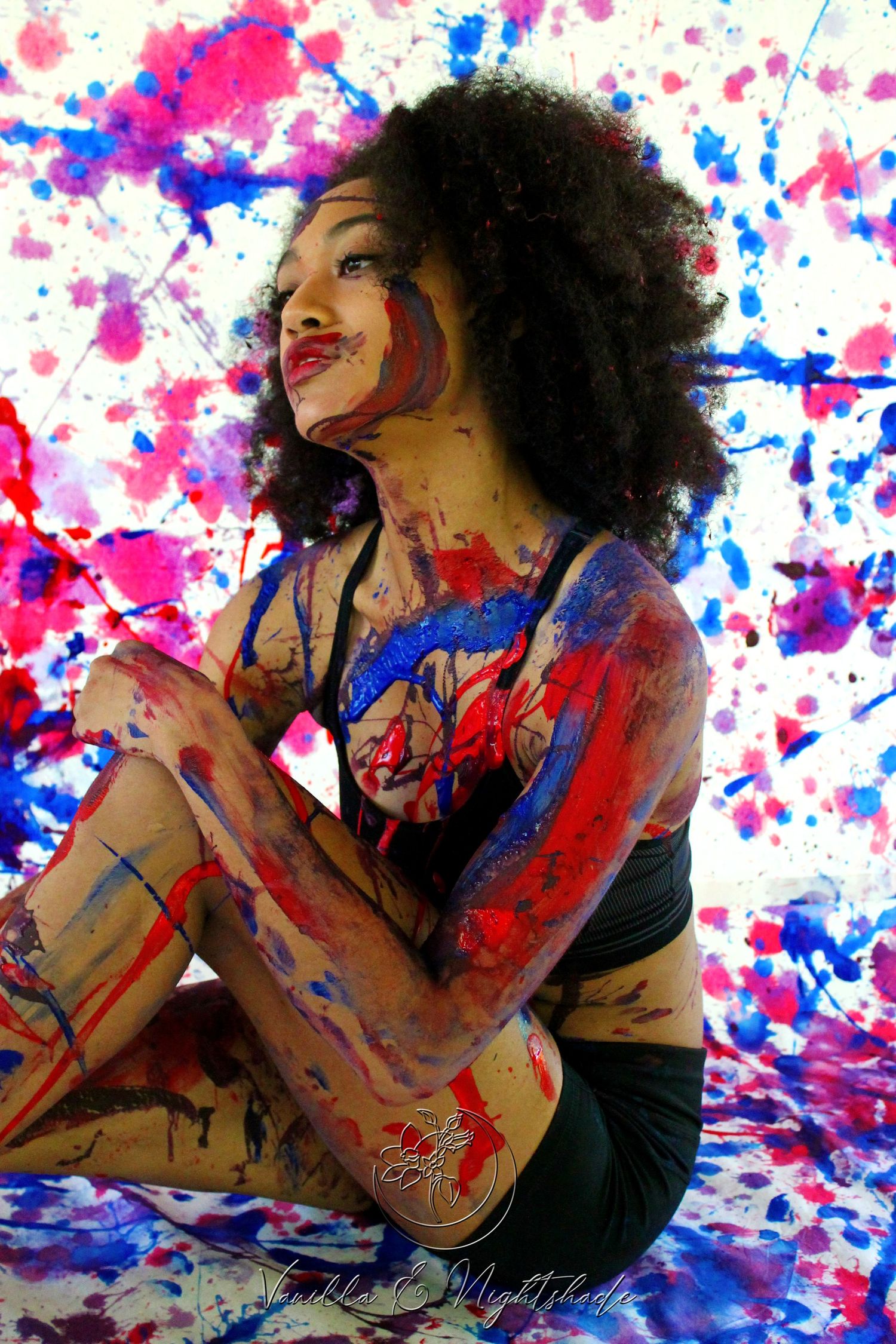 A woman with natural hair sitting against a colorful red, blue, and purple paint-splattered background.