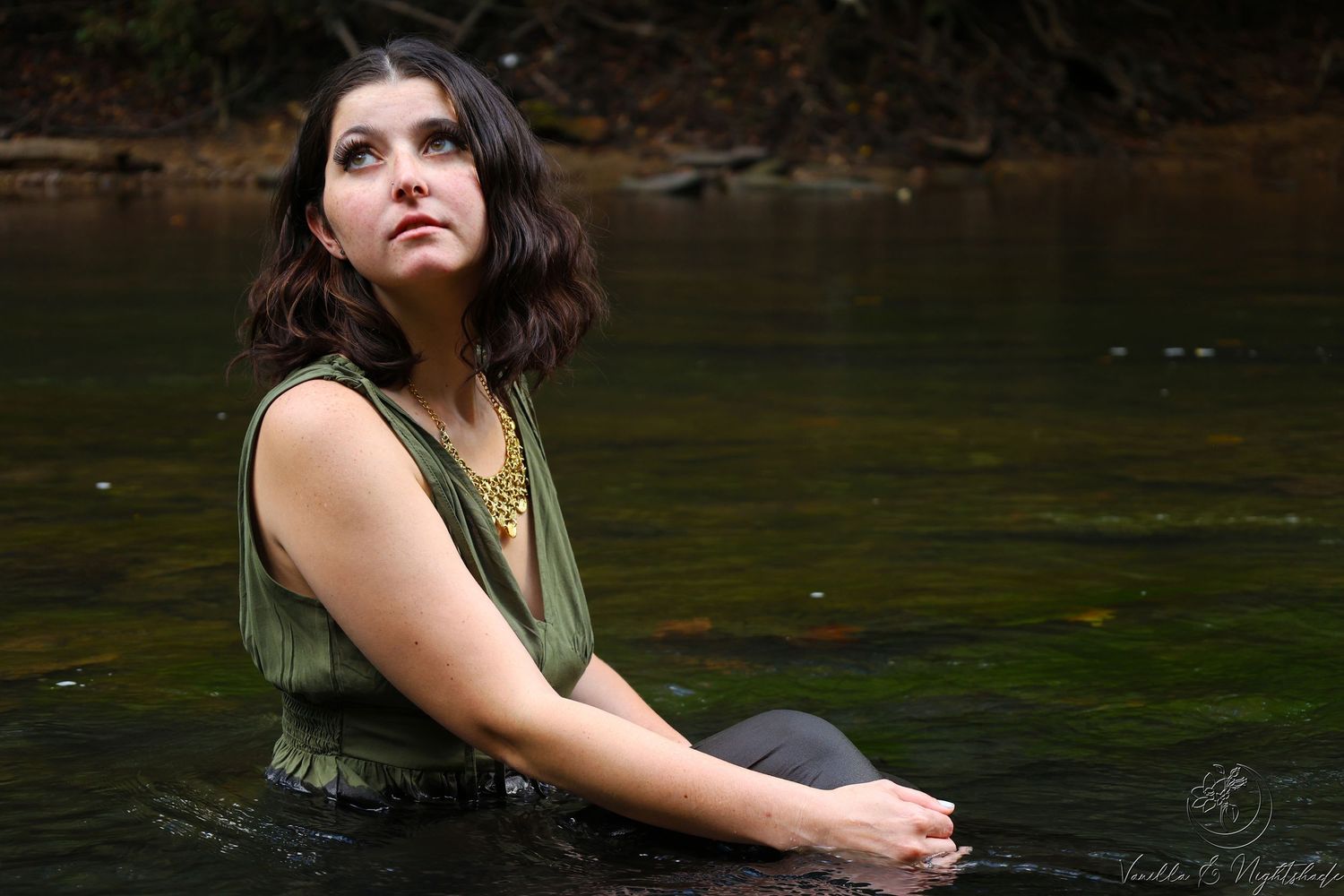Young brunette woman in olive top sits in shallow river water wearing gold necklace, looking up.