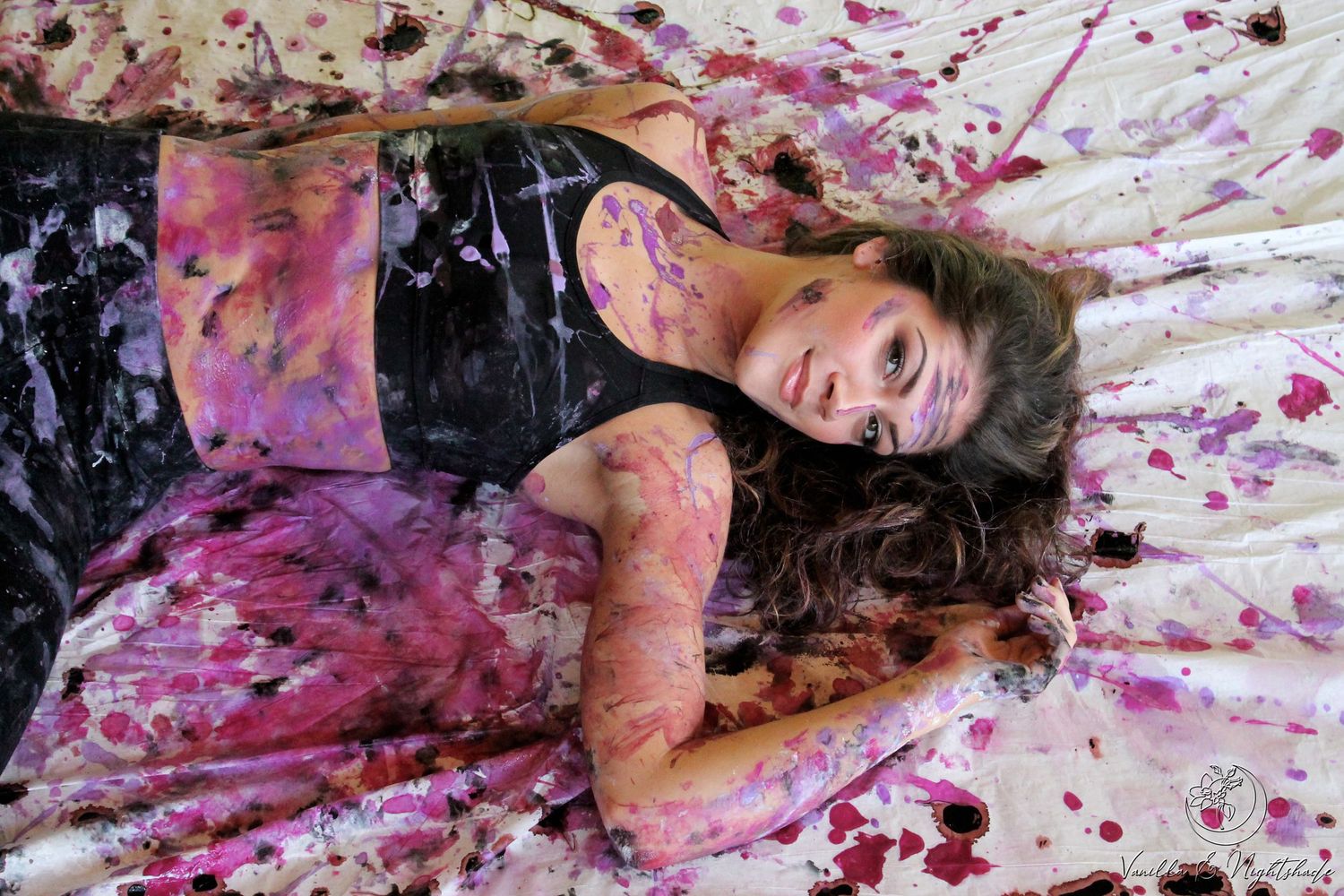 Woman lying on paint-splattered white surface covered in pink and black paint in artistic portrait.
