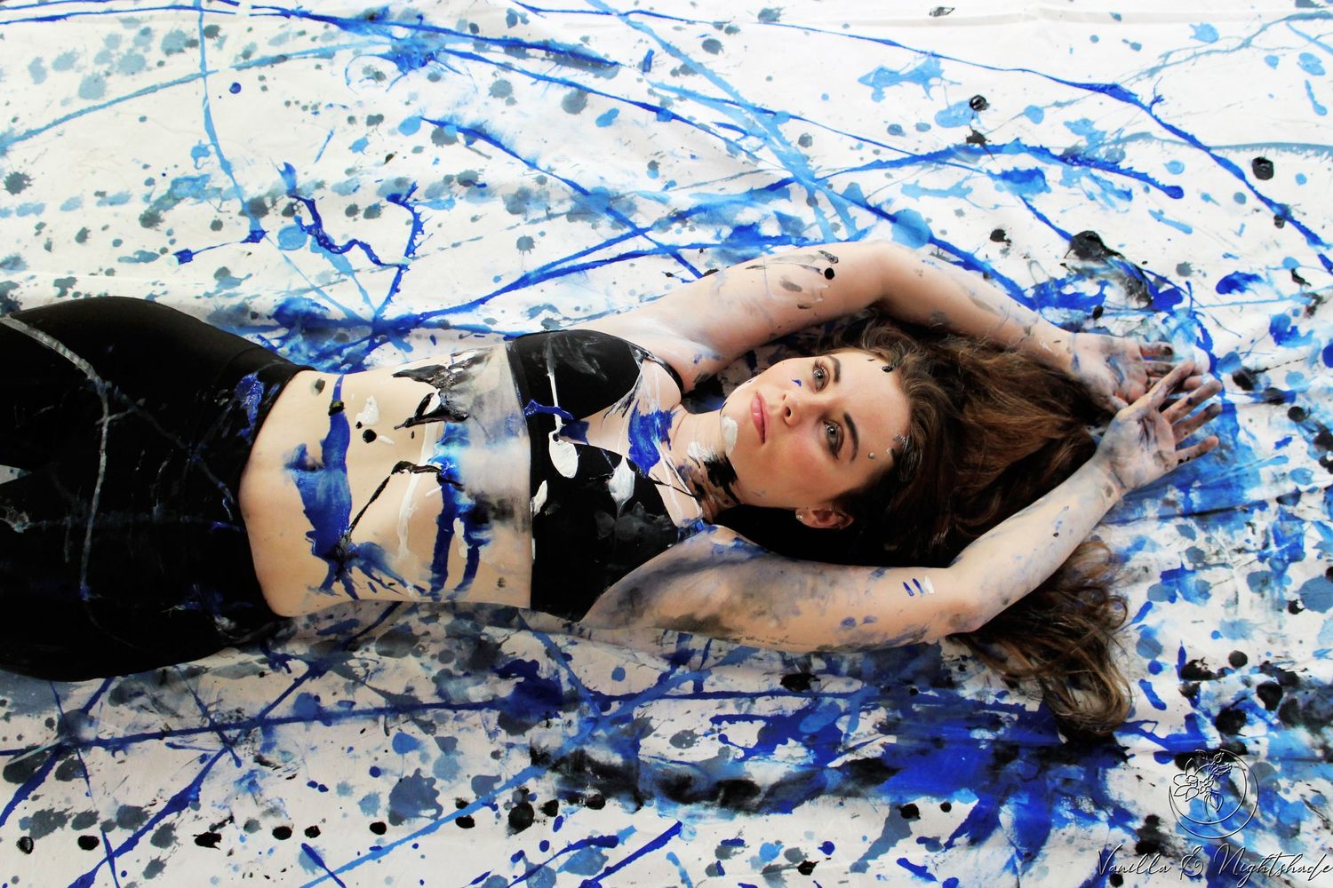 A young woman lying on a white surface covered in blue and black paint splatter artwork.