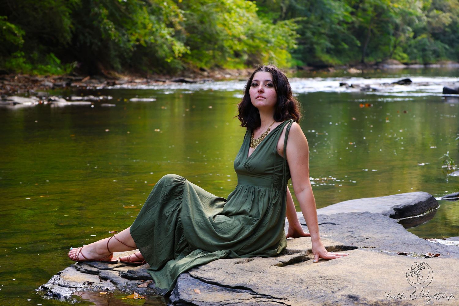 Brunette woman in elegant olive green dress poses on riverside rocks surrounded by forest greenery.