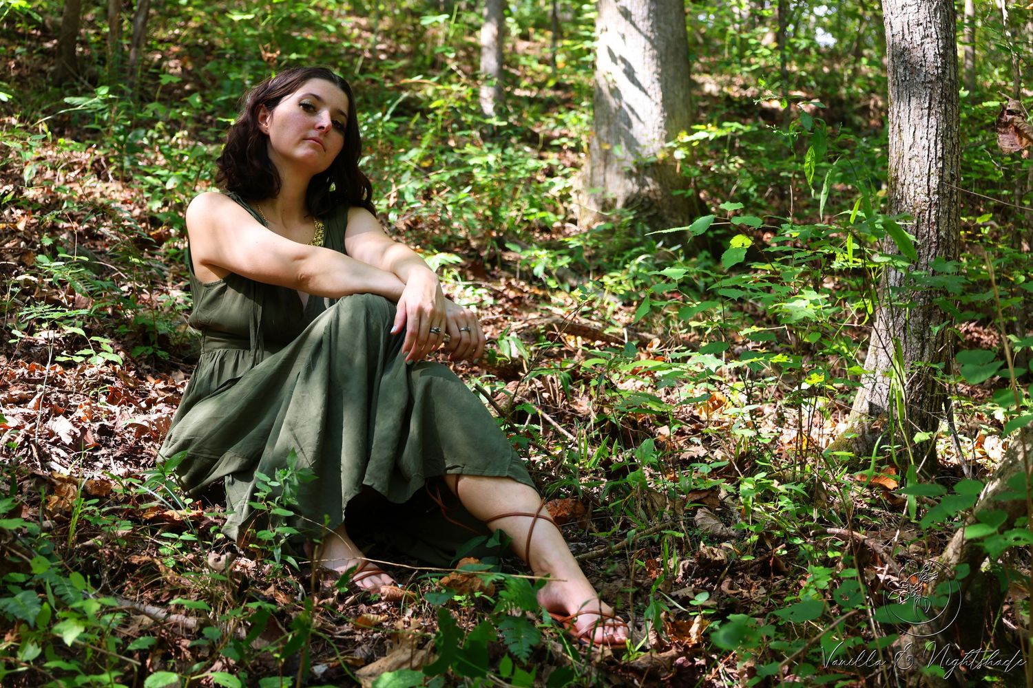 Woman in olive green dress sitting barefoot among green foliage on a forest floor.