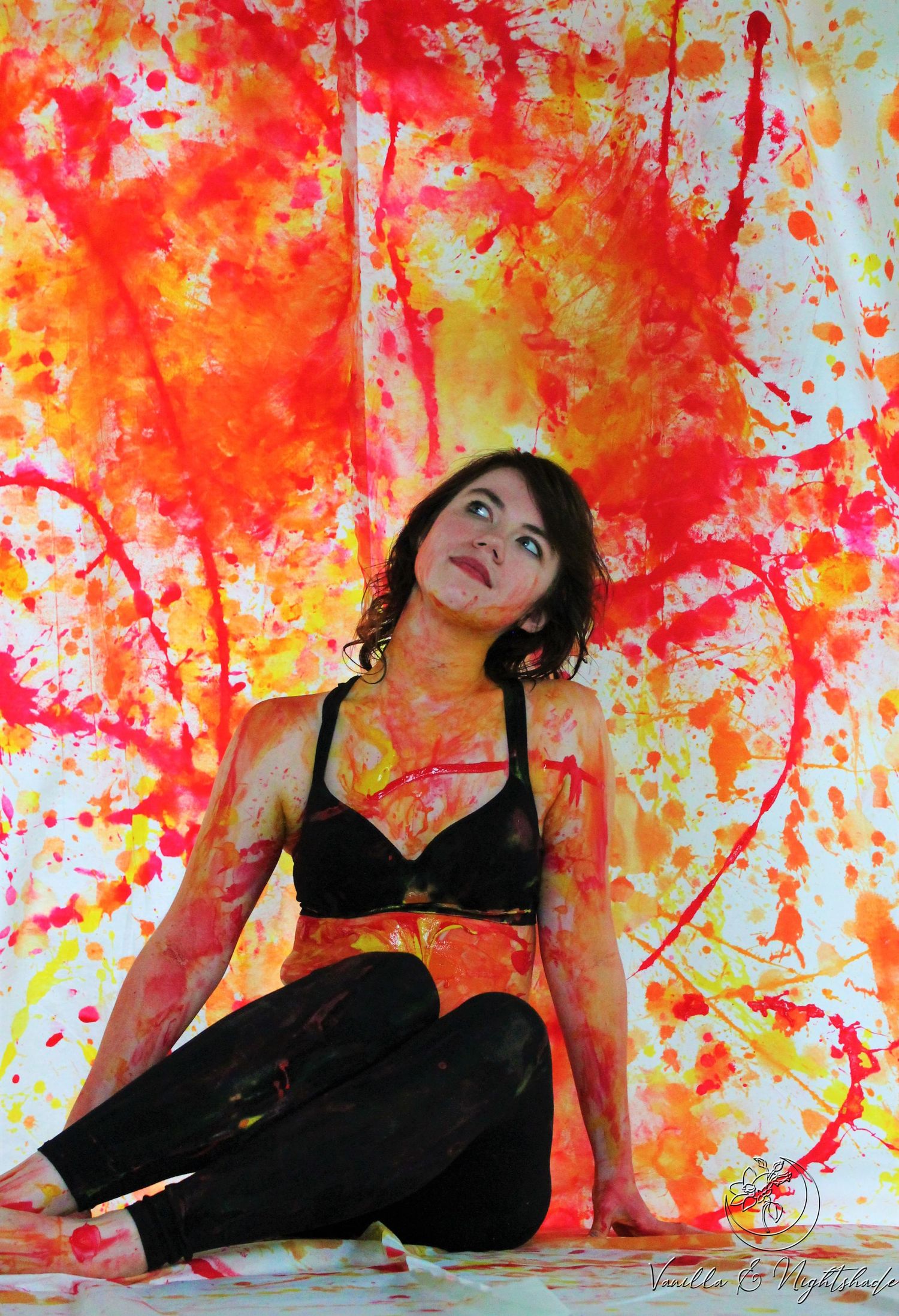 Woman covered in red, orange and yellow paint sits against vibrant splattered canvas backdrop.