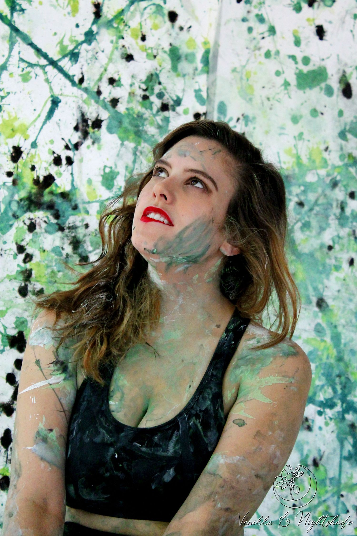 Woman with red lips covered in green and gray paint splatters looks up against splattered backdrop.