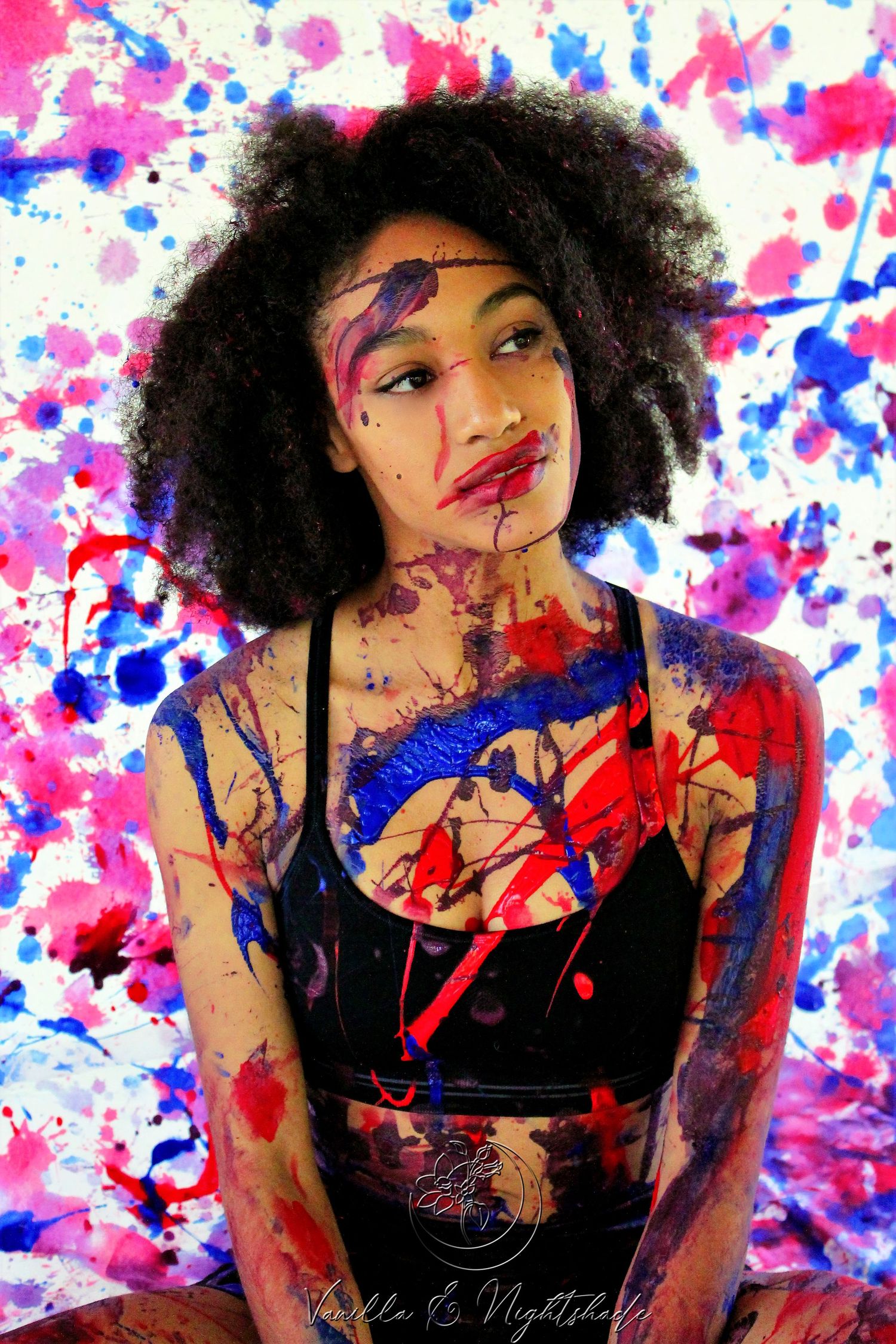 Woman with natural afro hair covered in colorful red, blue and purple paint splatters in artistic portrait.