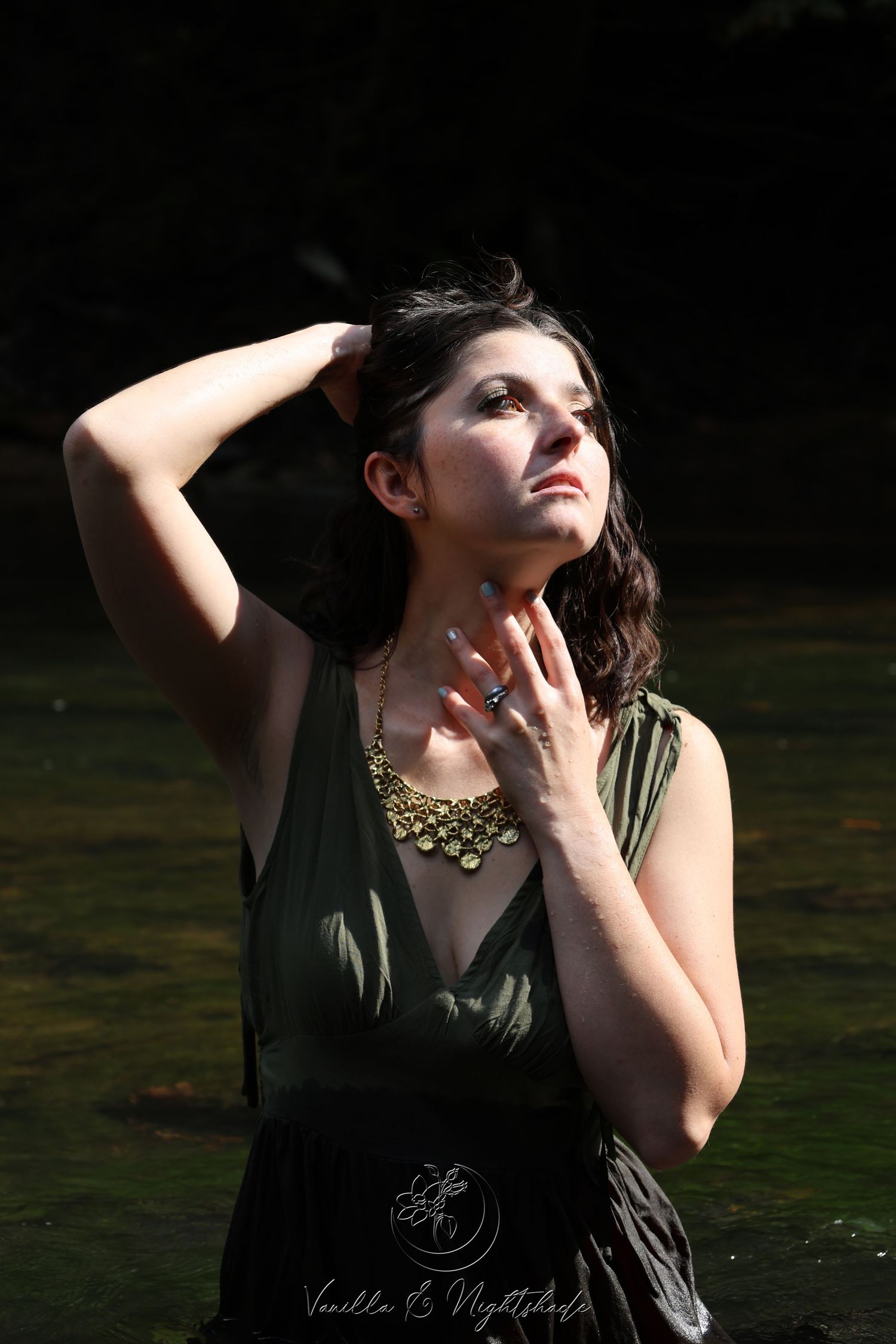 Woman in wet olive dress with gold necklace gazing upward in dramatic outdoor lighting.