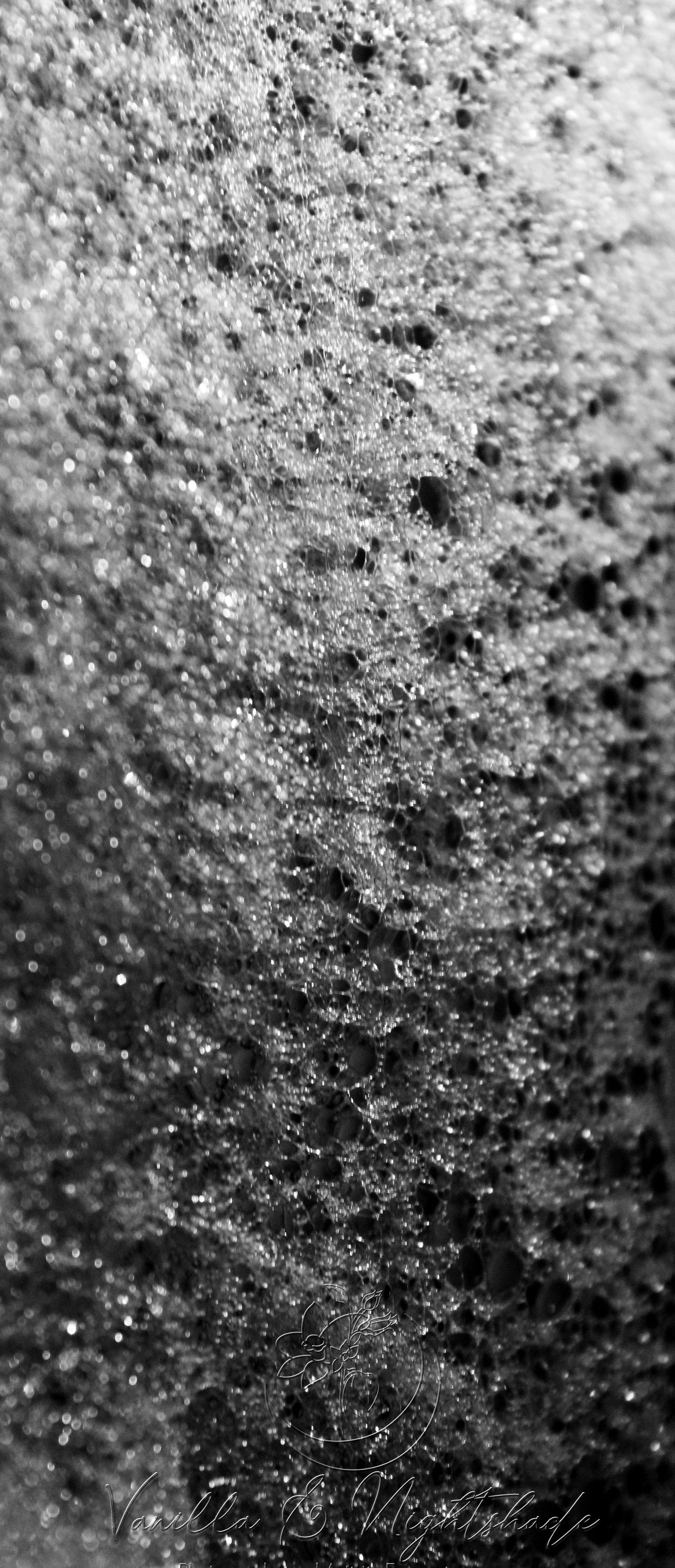 Close-up black and white macro photo of bubbling foam texture with sparkling droplets and dark pores.