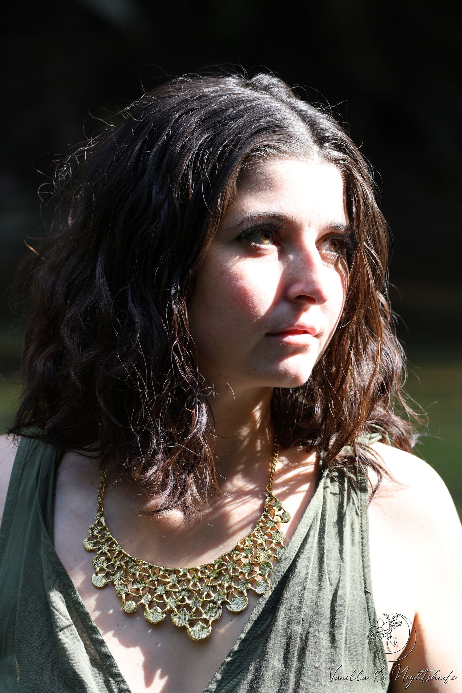 Dark-haired woman wearing gold statement necklace and olive top, sunlit portrait in natural setting.