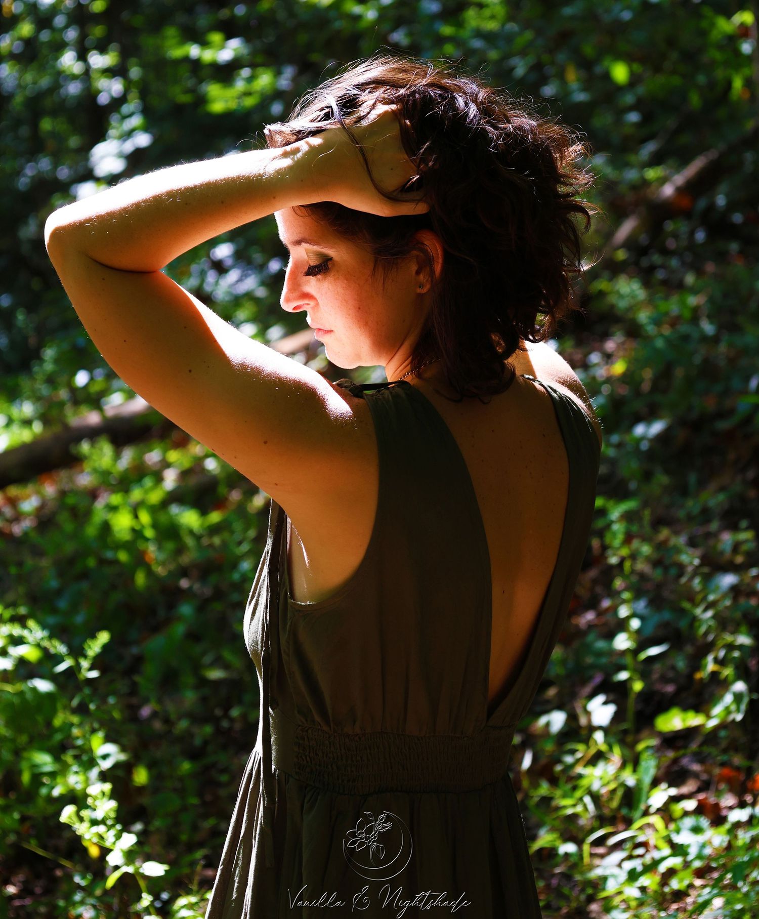Woman in olive backless dress shields eyes from dappled sunlight in a lush green forest.