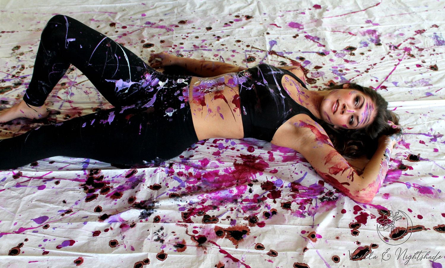 Woman lying on white paint-splattered surface covered in purple and red paint in artistic photo shoot.