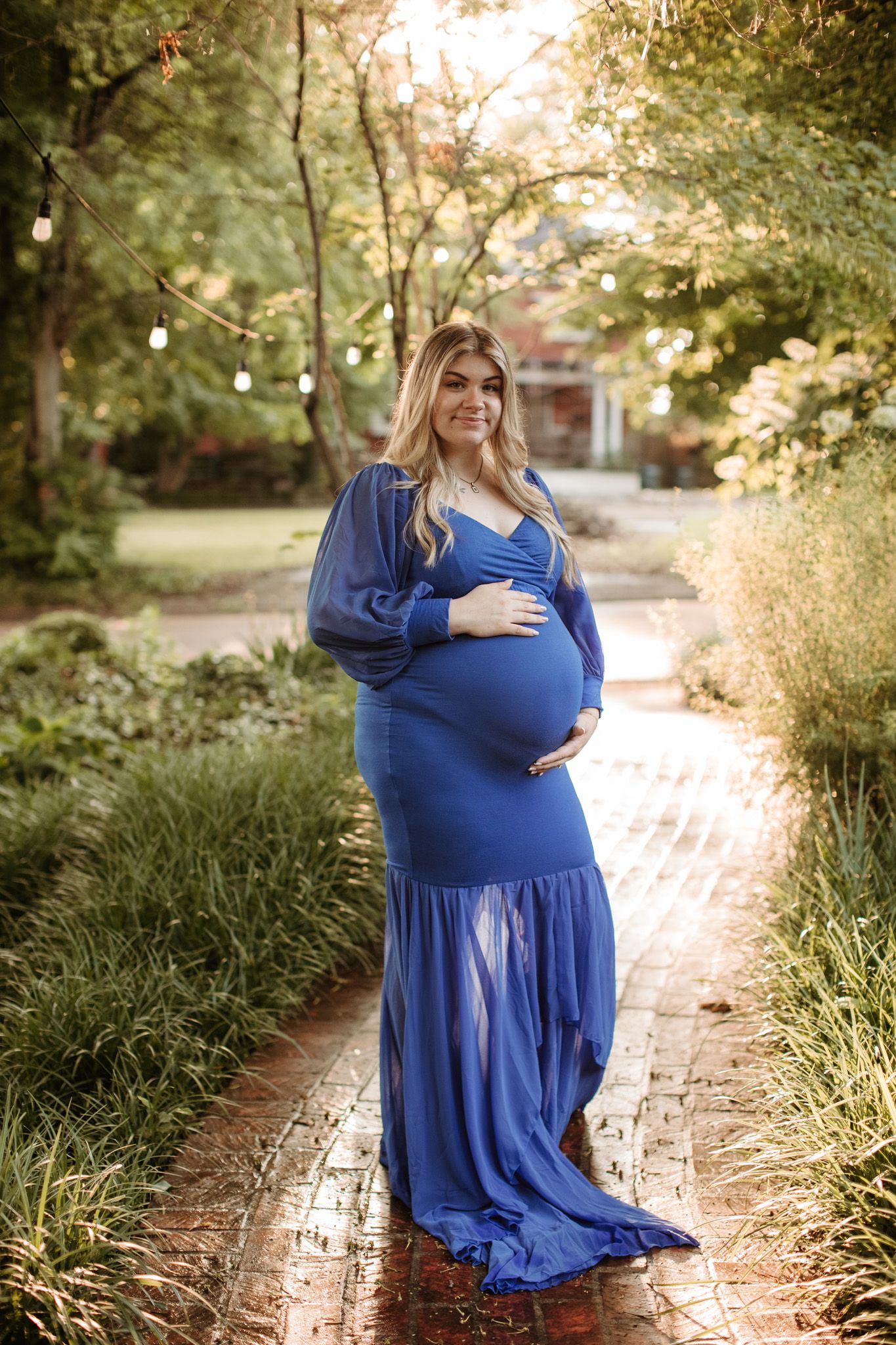 A pregnant woman in a flowing blue dress stands on a garden path during golden hour sunset.