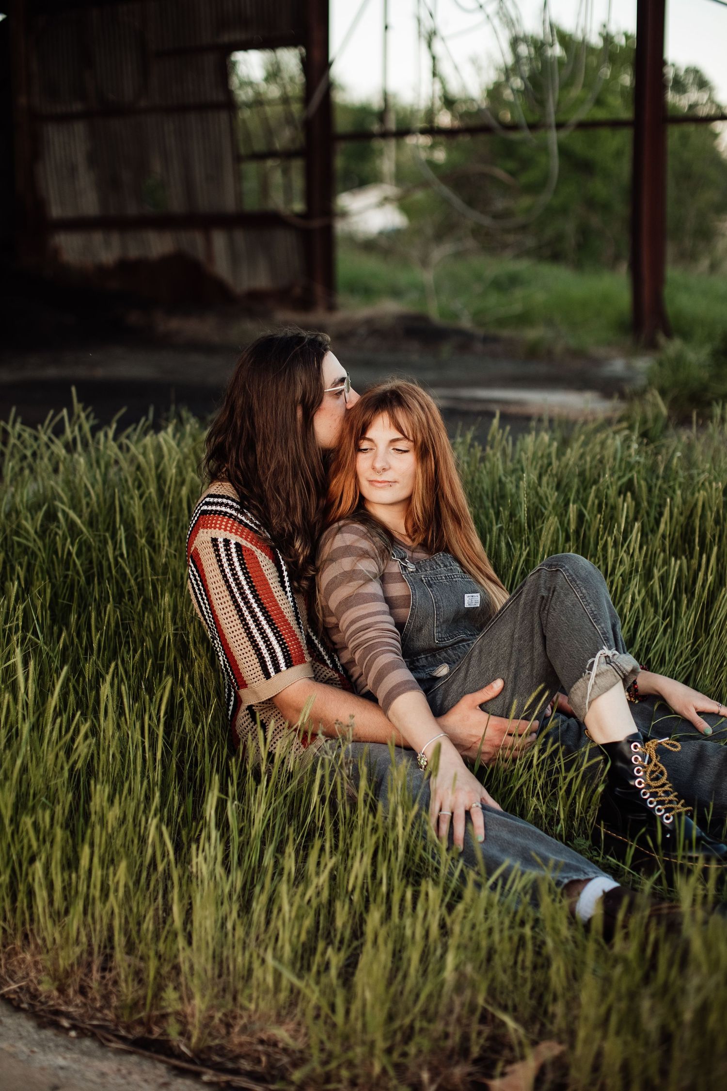 Two people in casual clothing sit together intimately in tall grass at sunset near an industrial structure.