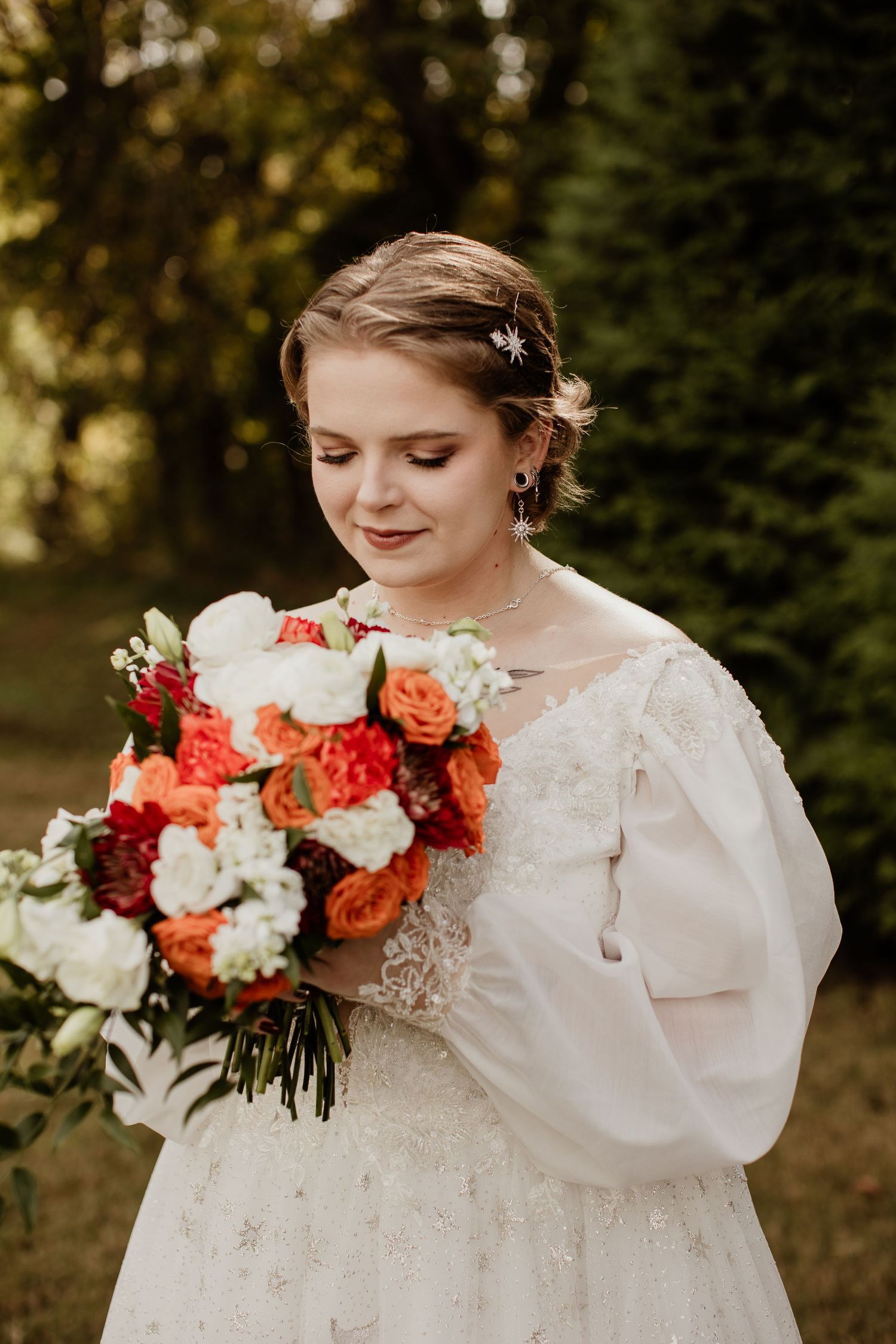 Bride in lace gown with puff sleeves holds vibrant orange and white bouquet outdoors, smiling softly.
