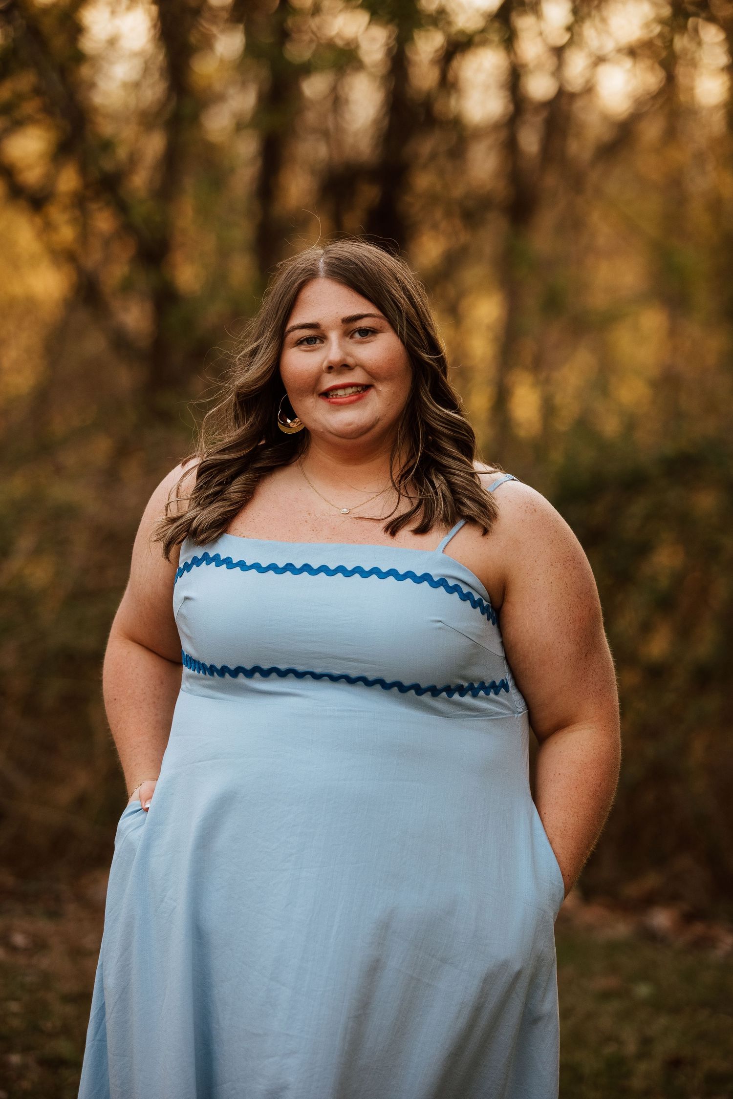 Young woman in light blue sundress with blue trim smiling outdoors in a wooded setting at golden hour.