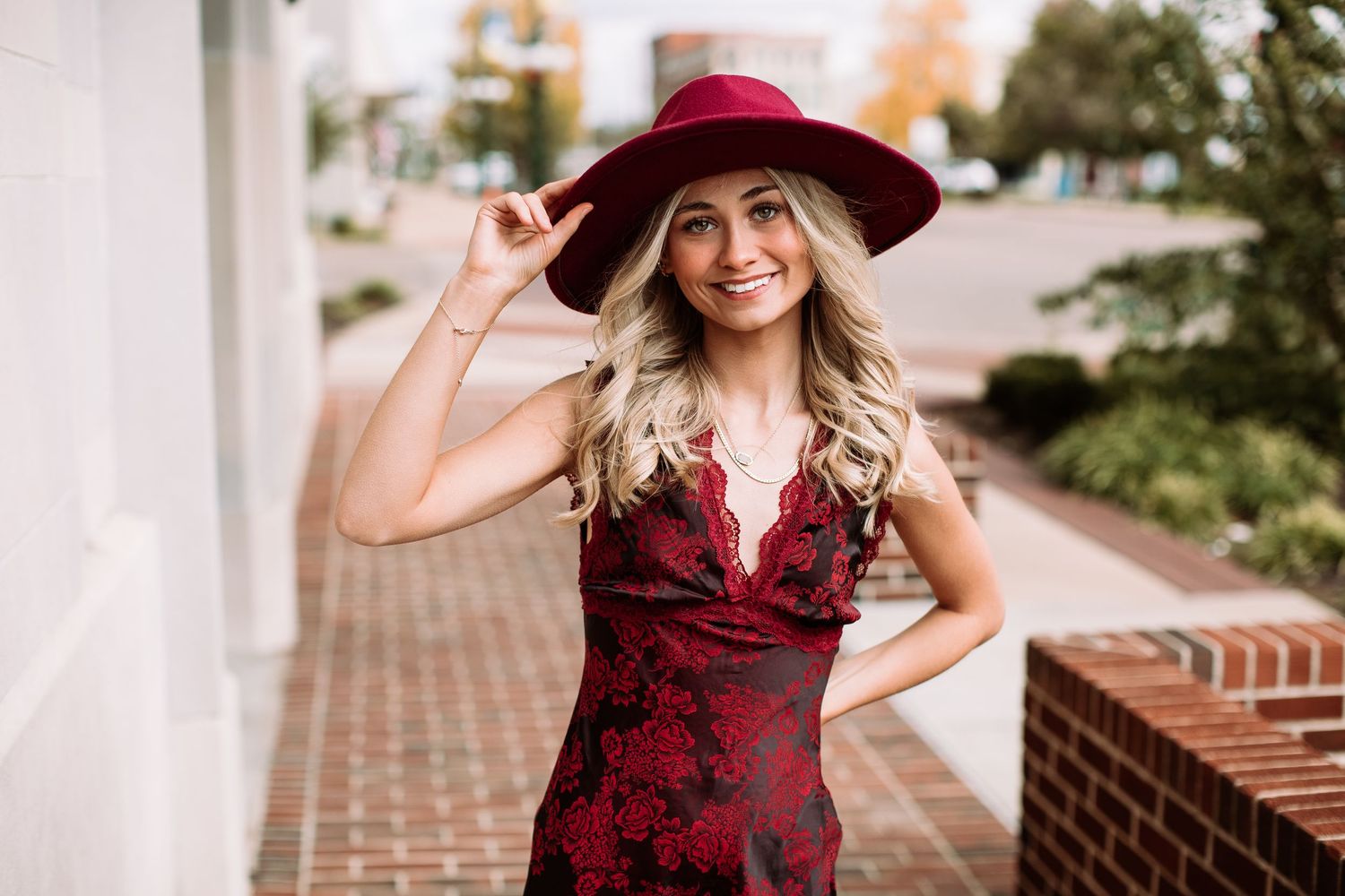 Stylish woman in burgundy dress and matching wide-brimmed hat poses on brick sidewalk with bright smile.