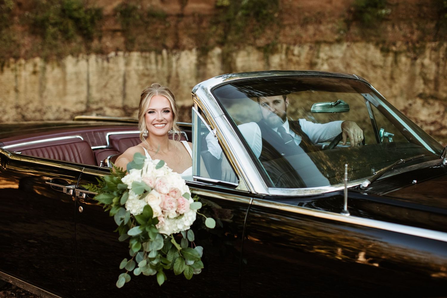 Elegant wedding party poses with vintage black car decorated with white floral arrangements and greenery.