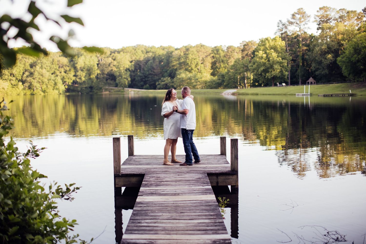 A romantic engagement photo session takes place on a wooden dock overlooking a serene lake during golden hour.