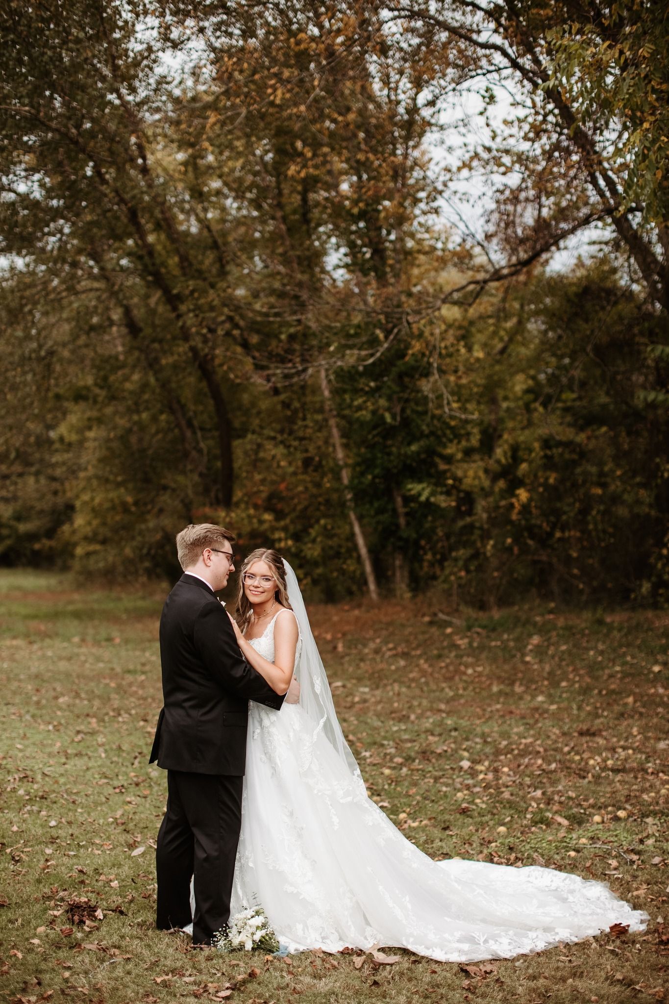 Bride and groom share a romantic moment in an autumn forest, her lace gown and veil trailing on the leaf-covered ground.